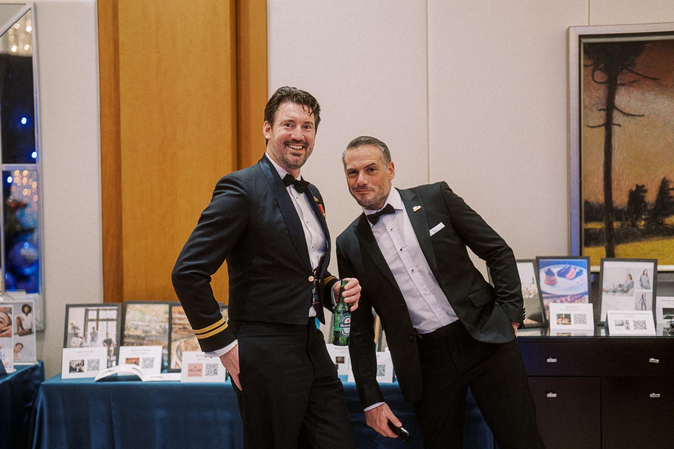 Two men in formal attire smiling at an event, standing in front of a display table with framed pictures and brochures.