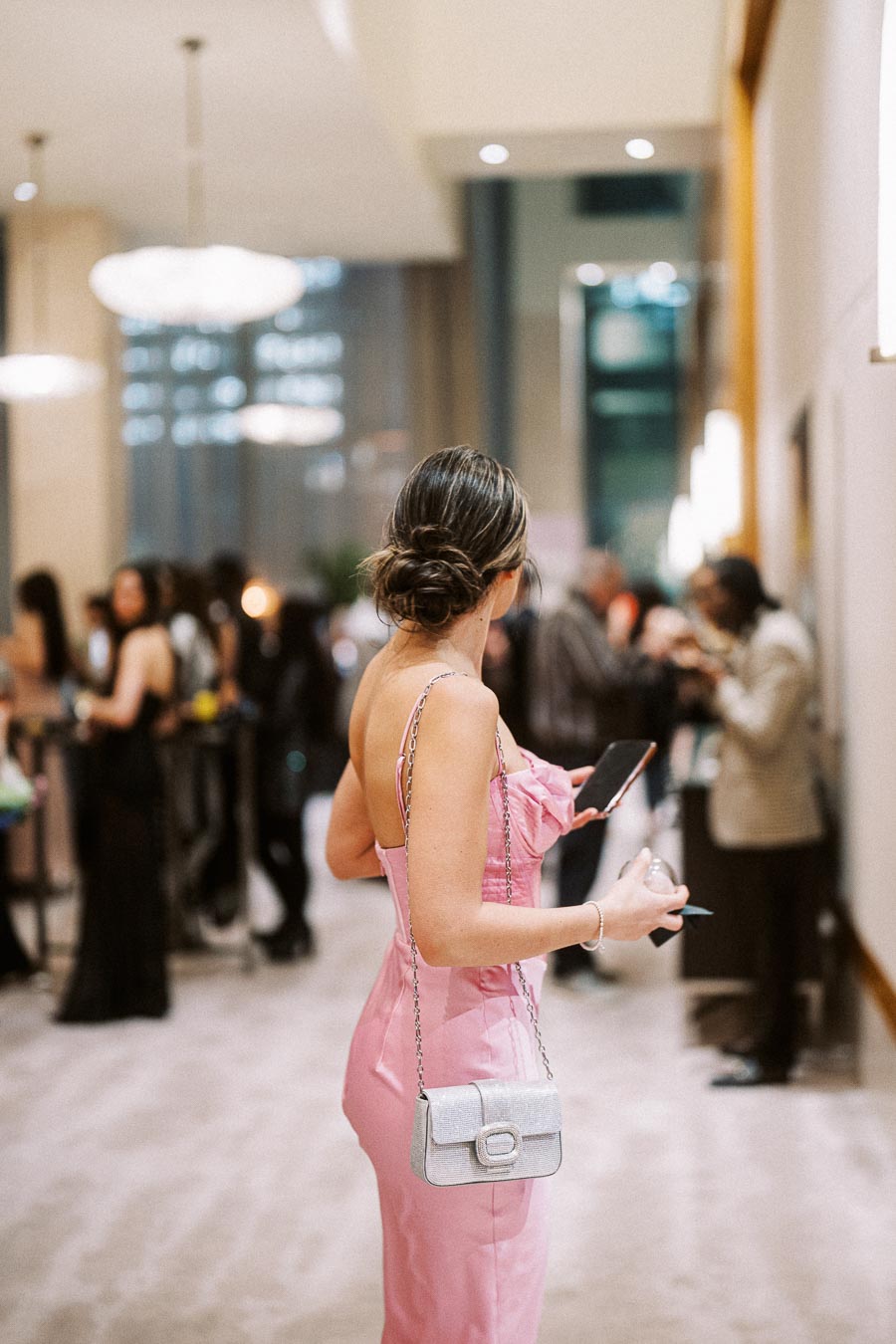 Woman in a pink dress holding a smartphone at a formal event, surrounded by elegantly dressed guests in a modern venue with soft lighting.