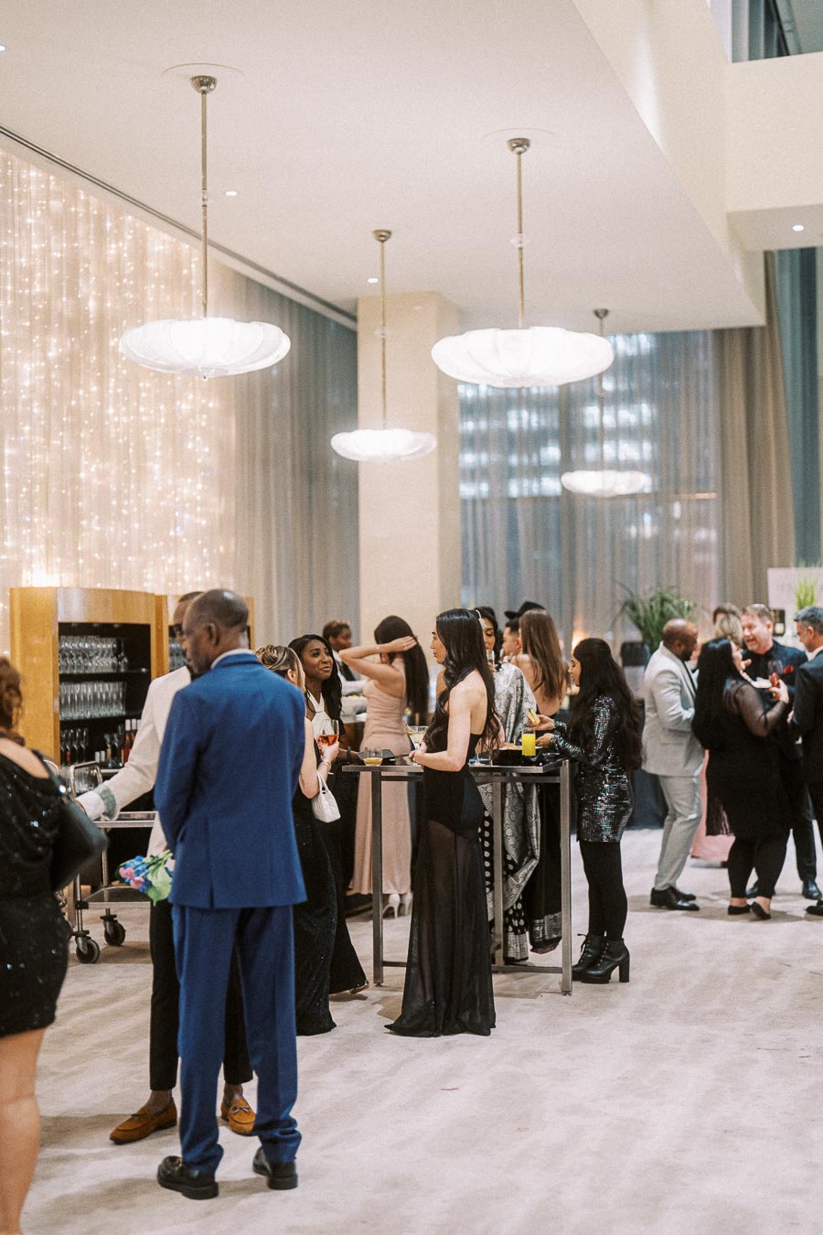 Elegant evening event with people mingling in a sophisticated venue, featuring modern lighting and decor. Guests in formal attire socialize around cocktail tables, creating a lively and upscale atmosphere.