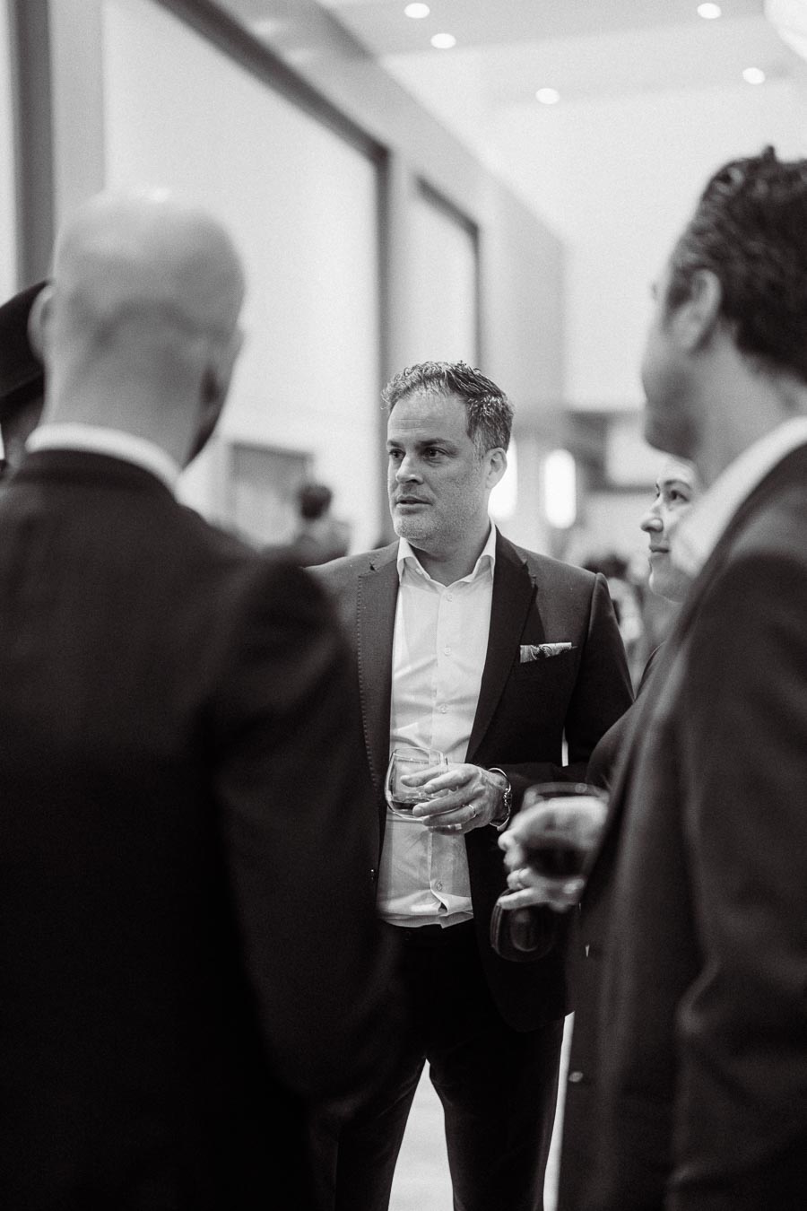 Black and white photo of a group of professional individuals in suits engaged in conversation, holding drinks, during a networking event in a modern indoor setting.