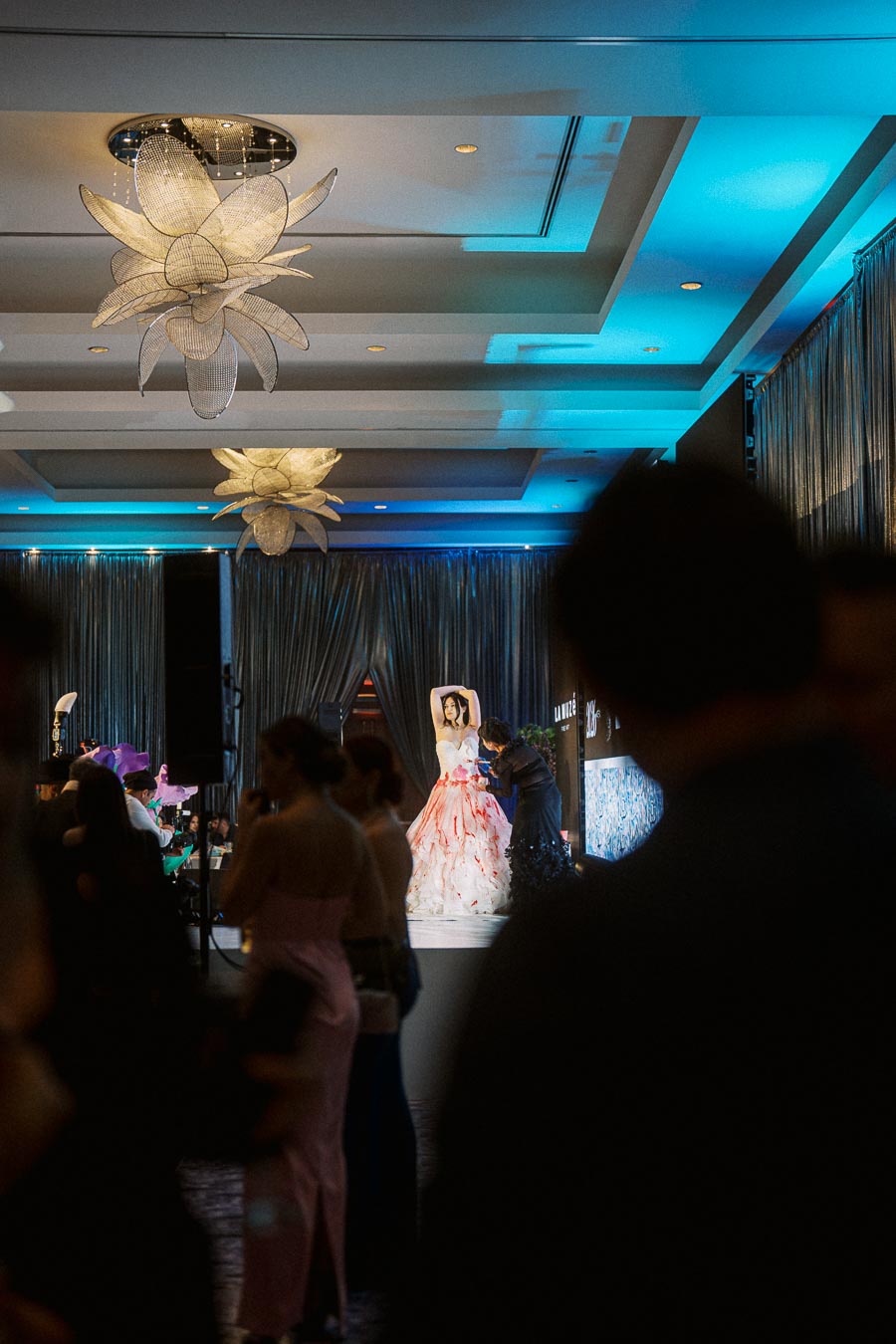 Elegant ballroom event with a woman in a striking white and red gown on stage, surrounded by a stylish crowd under artistic lighting fixtures and blue ambient lighting.