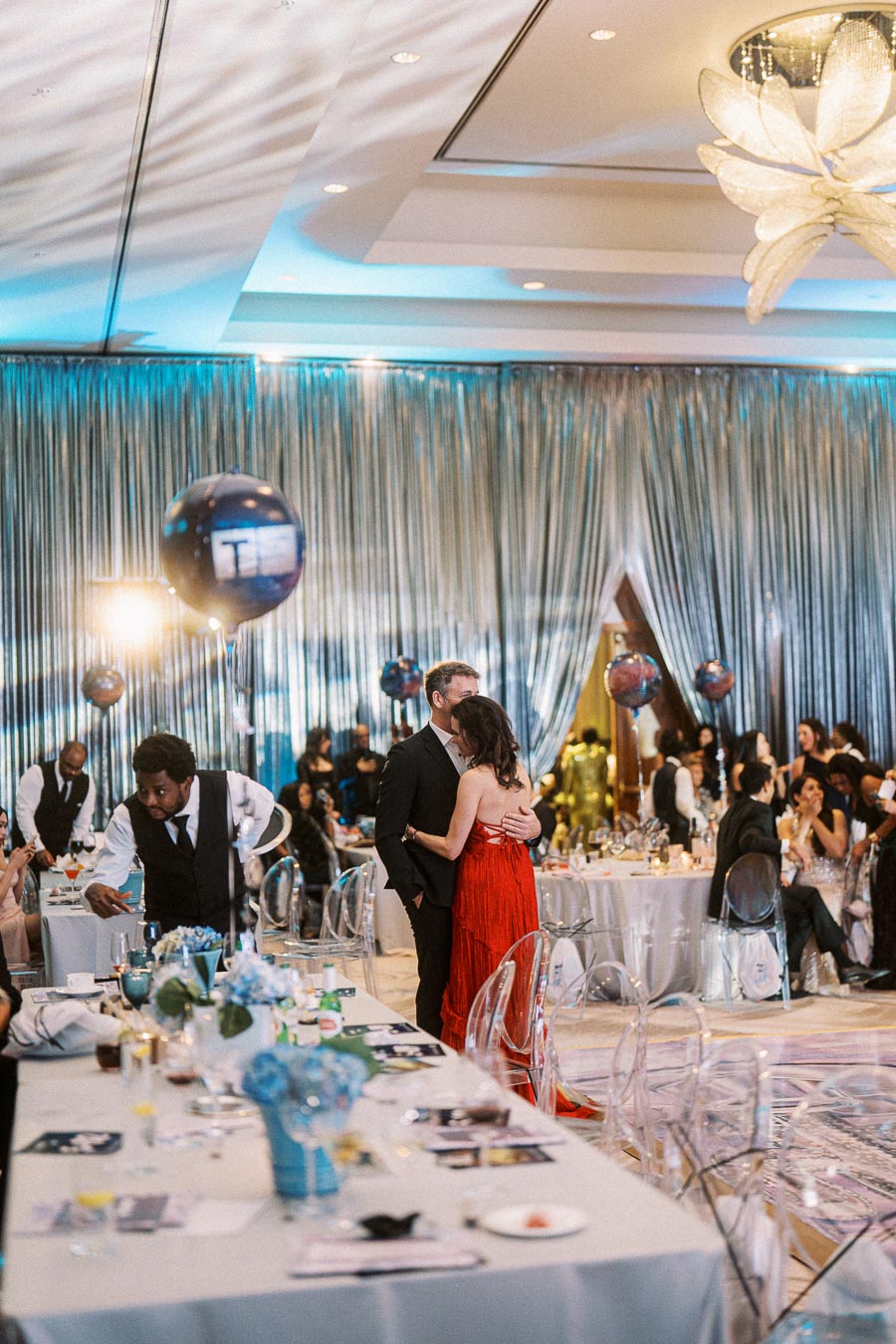 Elegant ballroom event with a couple enjoying a slow dance, surrounded by guests at decorated tables, featuring blue and silver drapery, balloons, and ambient lighting.