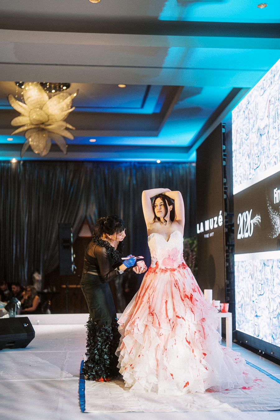 Fashion performance art featuring a model in a white gown being painted with bold red accents on stage, under dramatic lighting with an elegant chandelier, capturing an avant-garde art experience.