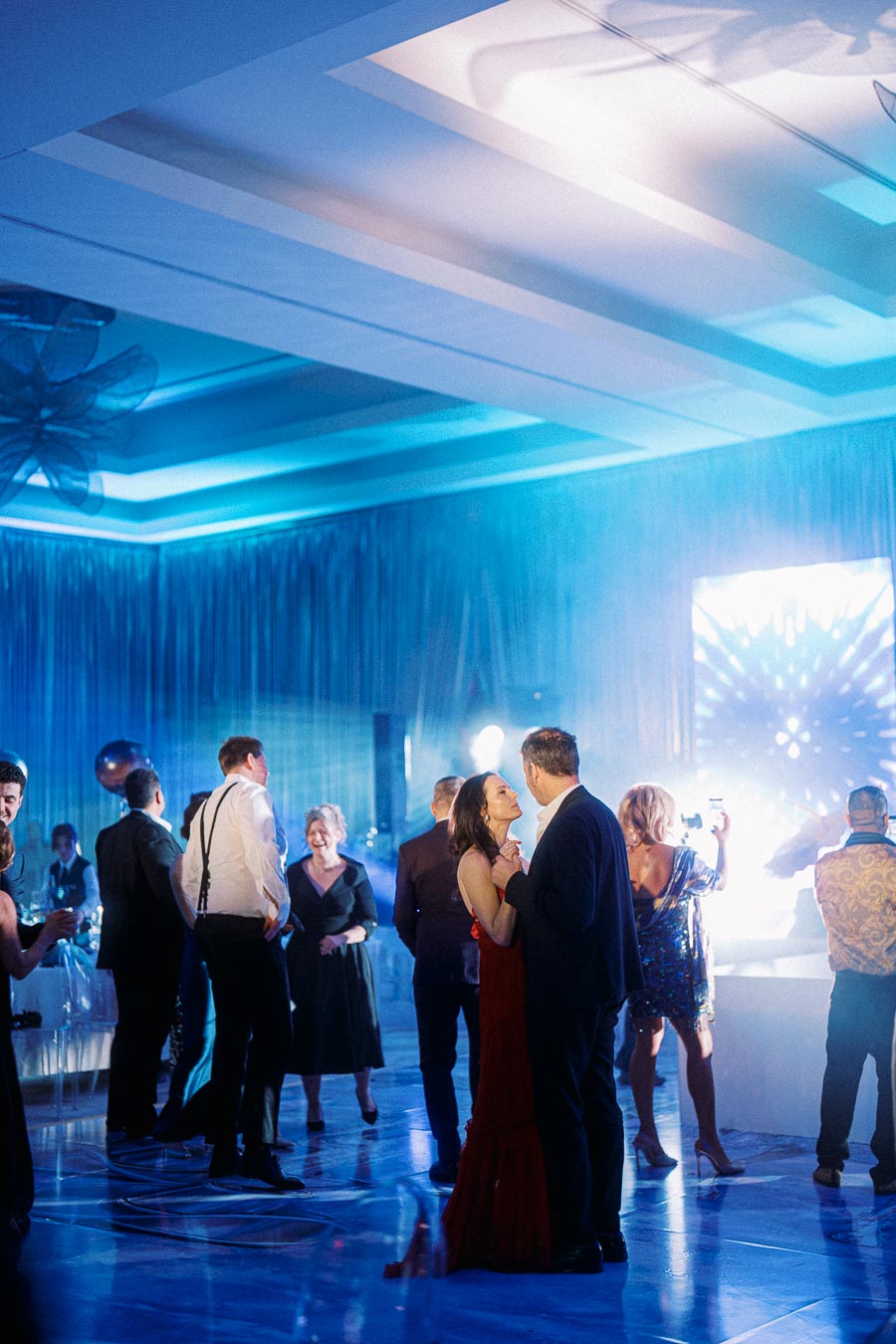 Couples elegantly dressed dancing at a vibrant evening party with blue ambient lighting and a lively atmosphere.