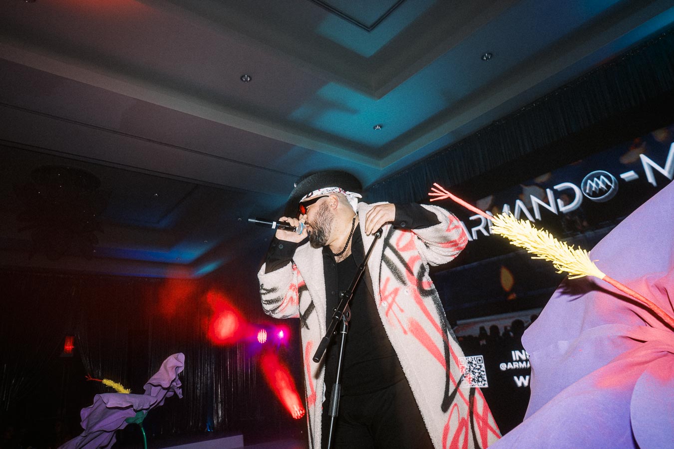 Performer on stage wearing a patterned coat and hat, singing into a microphone at a vibrant indoor event with colorful lighting effects.