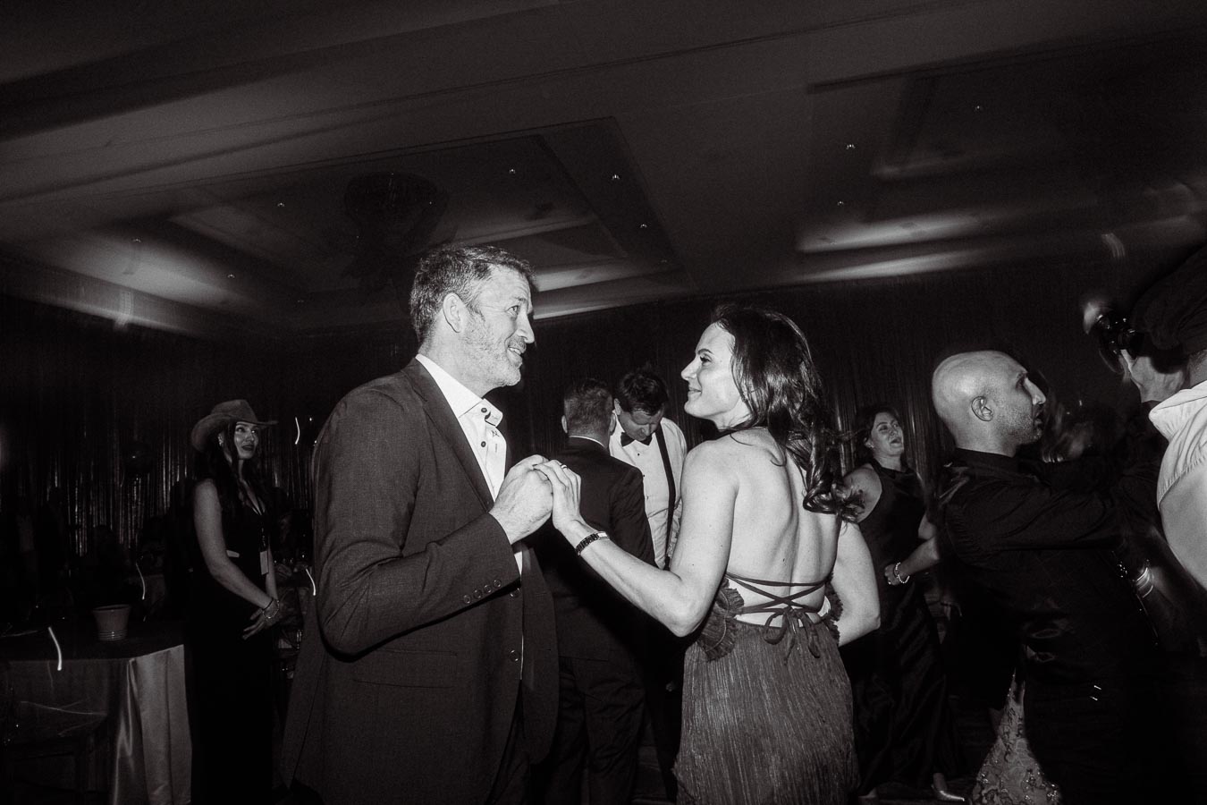 A couple dancing at a formal event with attendees enjoying the atmosphere, capturing the joyous and celebratory mood of the evening in black and white photography.