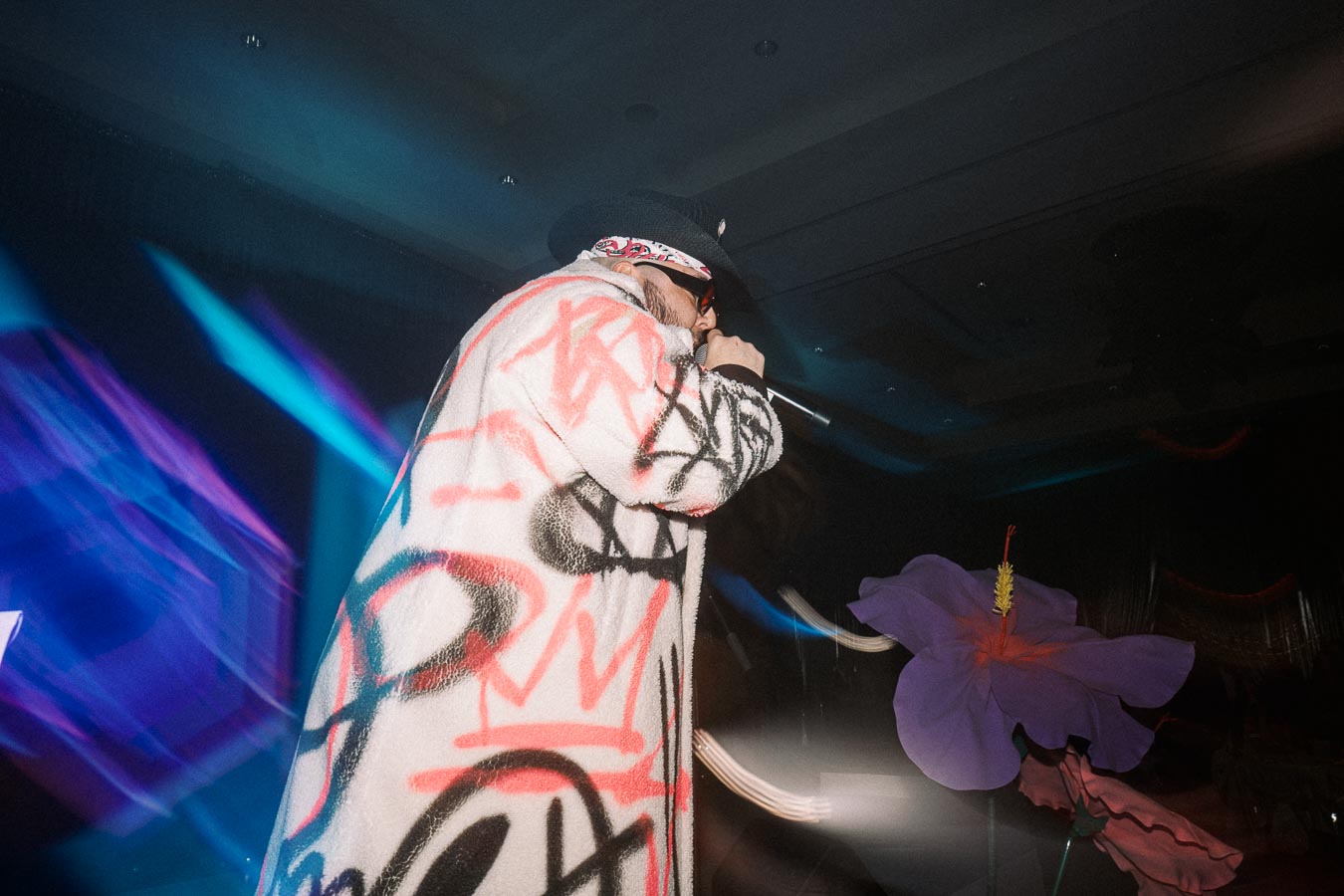 Live performance by an artist wearing a graffiti-patterned coat and black hat, singing into a microphone on a dimly lit stage with colorful lights and large artificial flowers in the background.