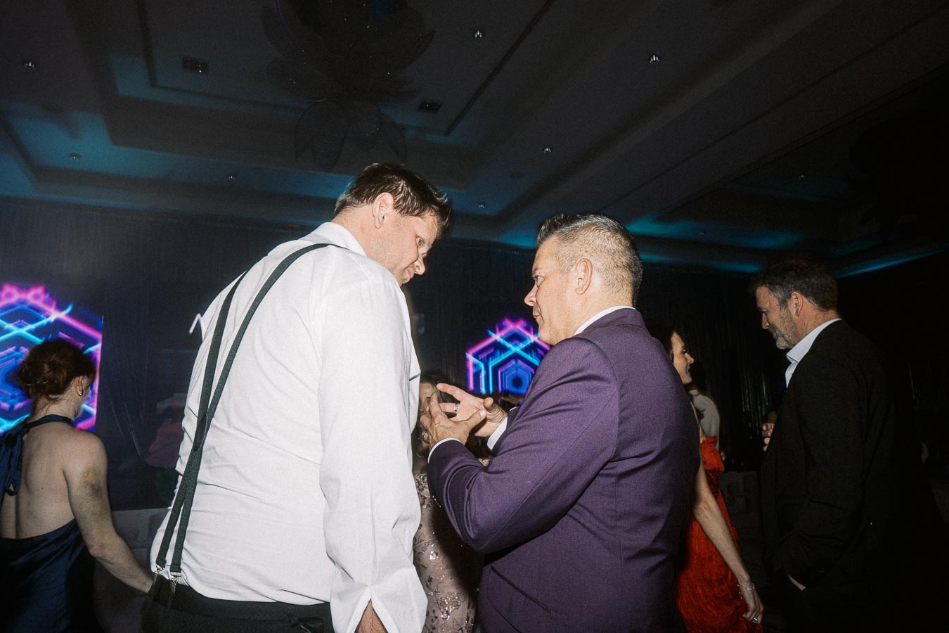 A group of people in formal attire engaged in conversation at an event with colorful geometric neon lights in the background.