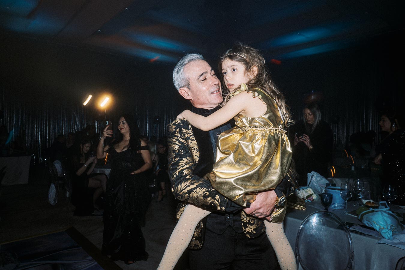 A man in a decorative suit holding a young girl in a gold dress at an indoor event, with people seated and taking photos in the background.