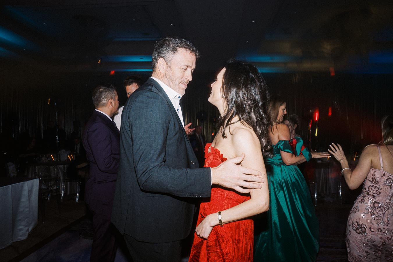 A couple dancing at a glamorous event, with guests socializing in an elegantly lit venue.