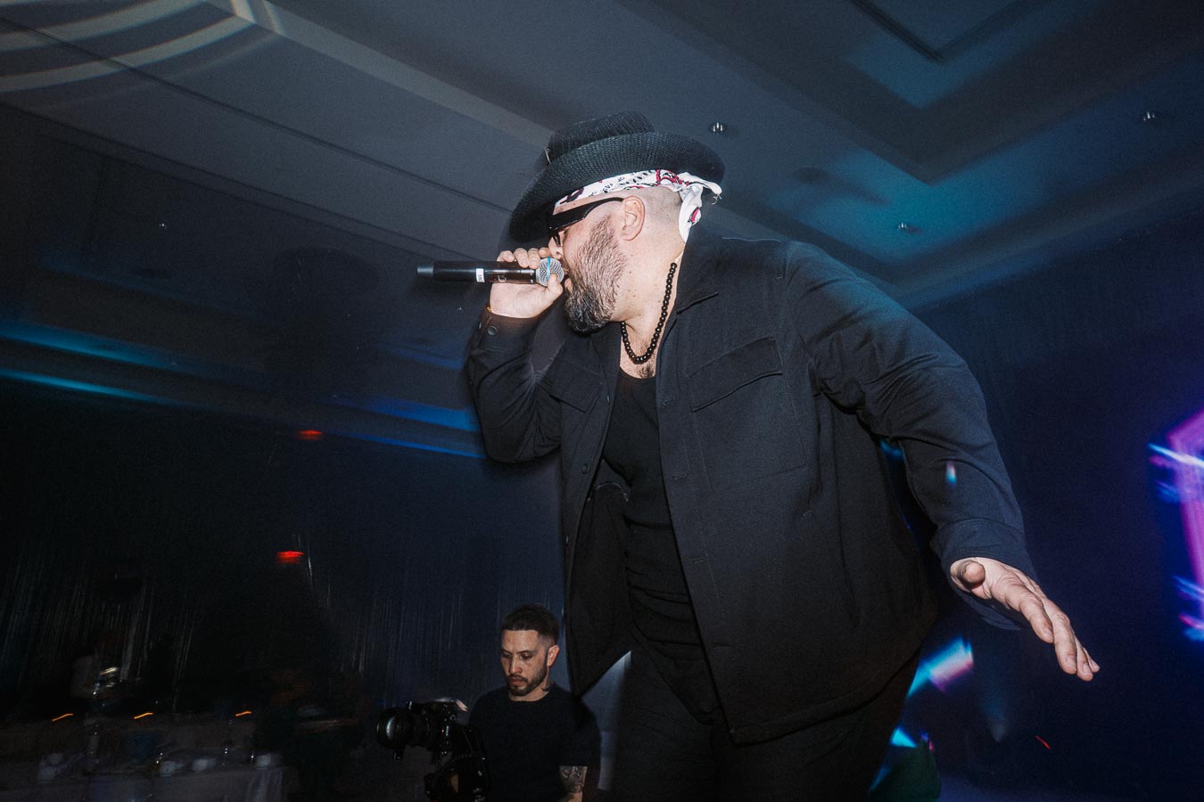 Musician performing on stage with microphone, wearing a black outfit and hat, in front of a lively audience and vibrant lighting.