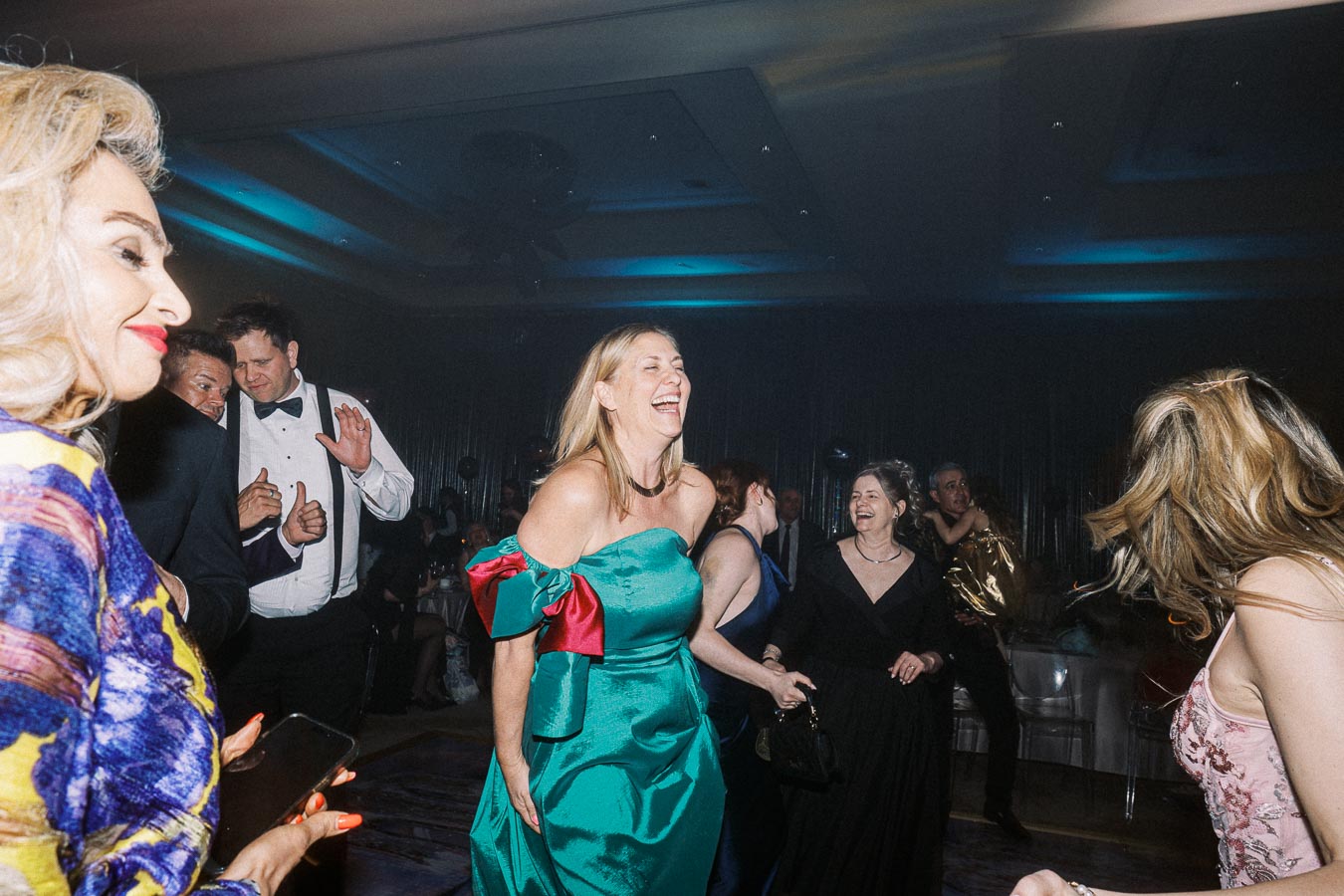 A lively group of people dancing and enjoying a formal event, showcasing elegant dresses and tuxedos, in a dimly lit ballroom with vibrant blue and gold lighting.