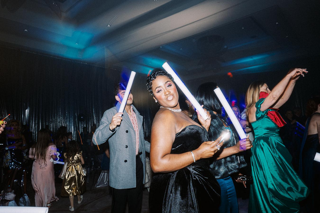 A lively group of people enjoying a party, holding glowing light sticks. A woman in a stylish black dress poses with a playful expression, surrounded by an energetic crowd under dim party lights.