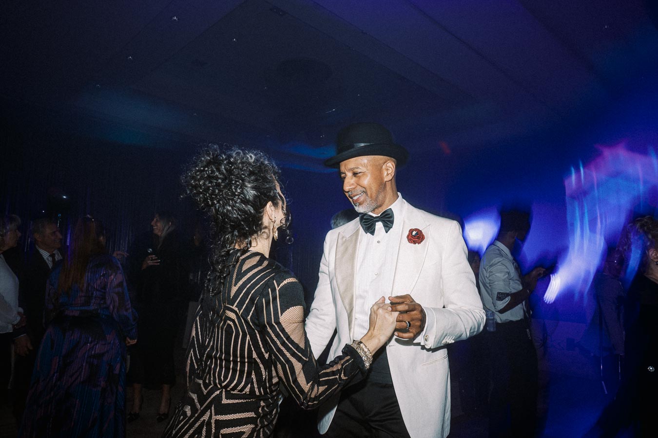 A couple elegantly dressed dances at a lively evening event, with the man in a white suit and black hat. The venue is dimly lit with colorful lights creating an energetic atmosphere. Guests in the background add to the festive mood.