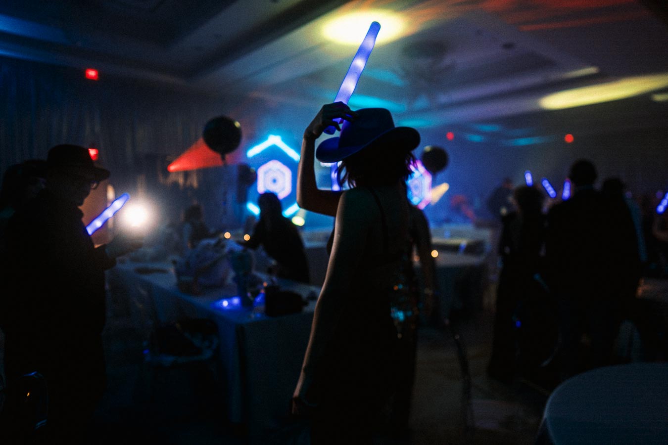 Person holding a glowing light stick in a dimly lit party setting with colorful lighting and silhouettes of people.