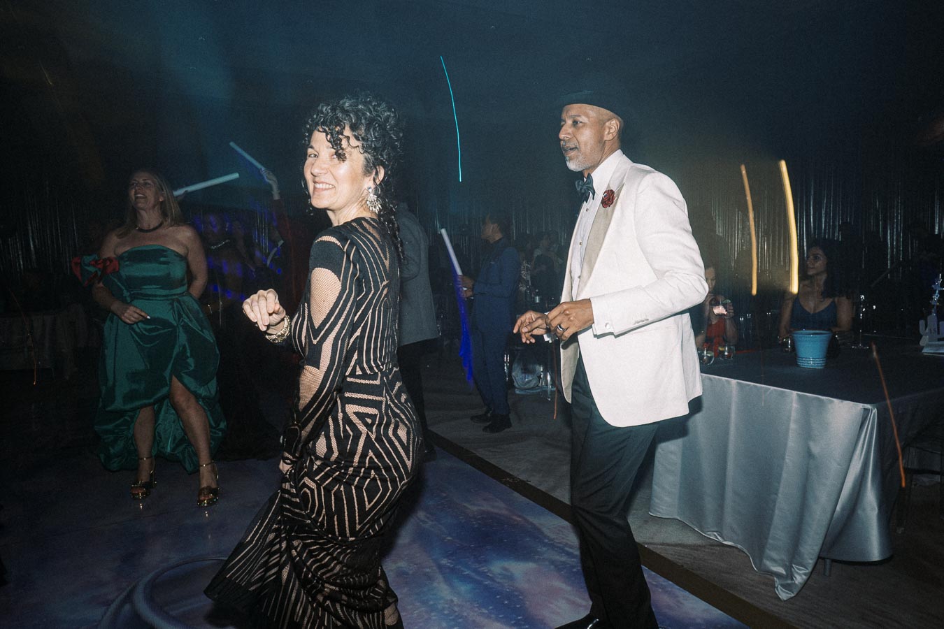 A group of elegantly dressed people dancing joyfully at a celebration or party event, with a woman in a patterned black dress and a man in a white suit jacket in the foreground, creating a lively and festive atmosphere.