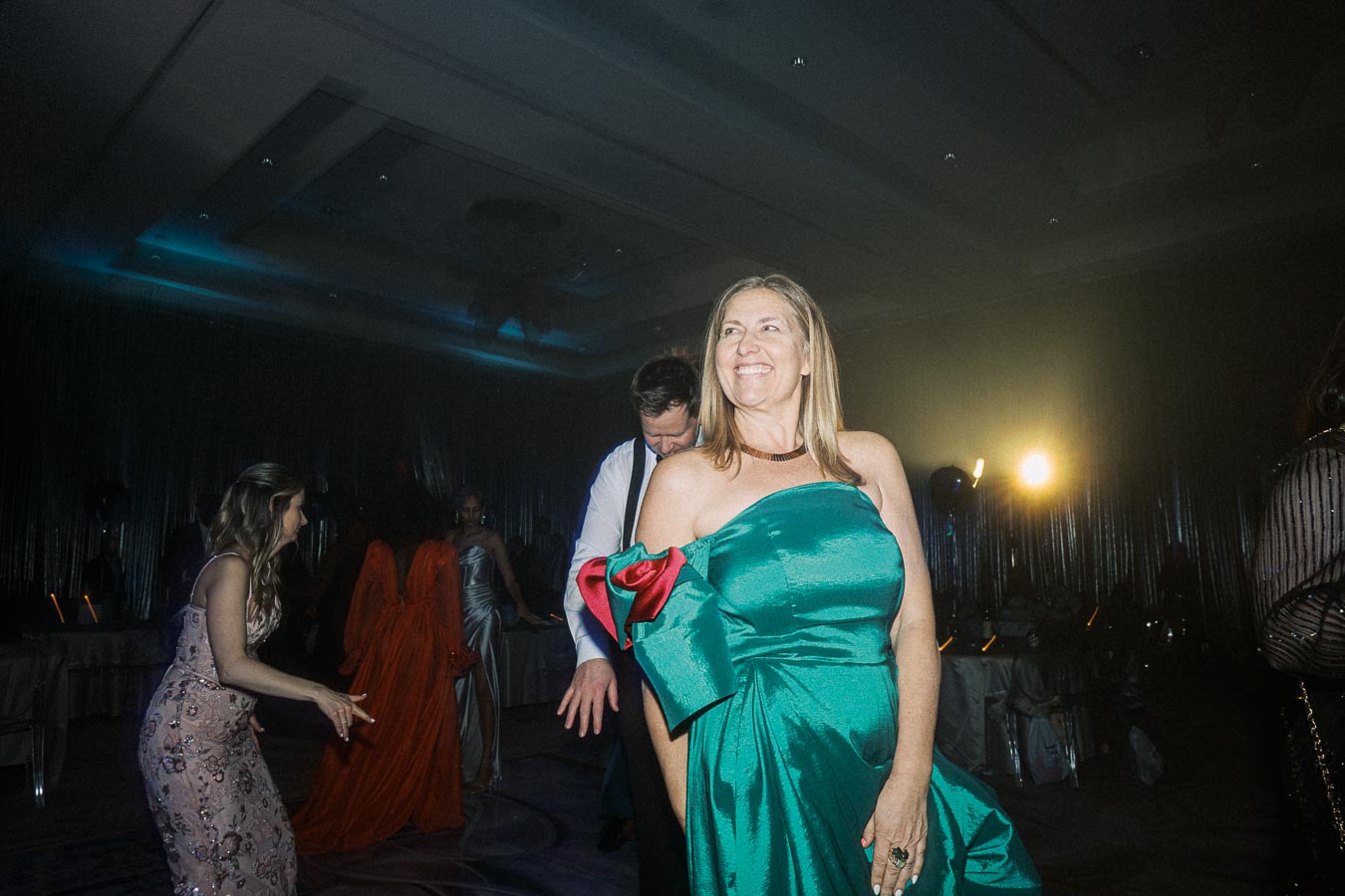 A woman in a vibrant green dress smiling at a lively event with dancing guests in dim lighting.