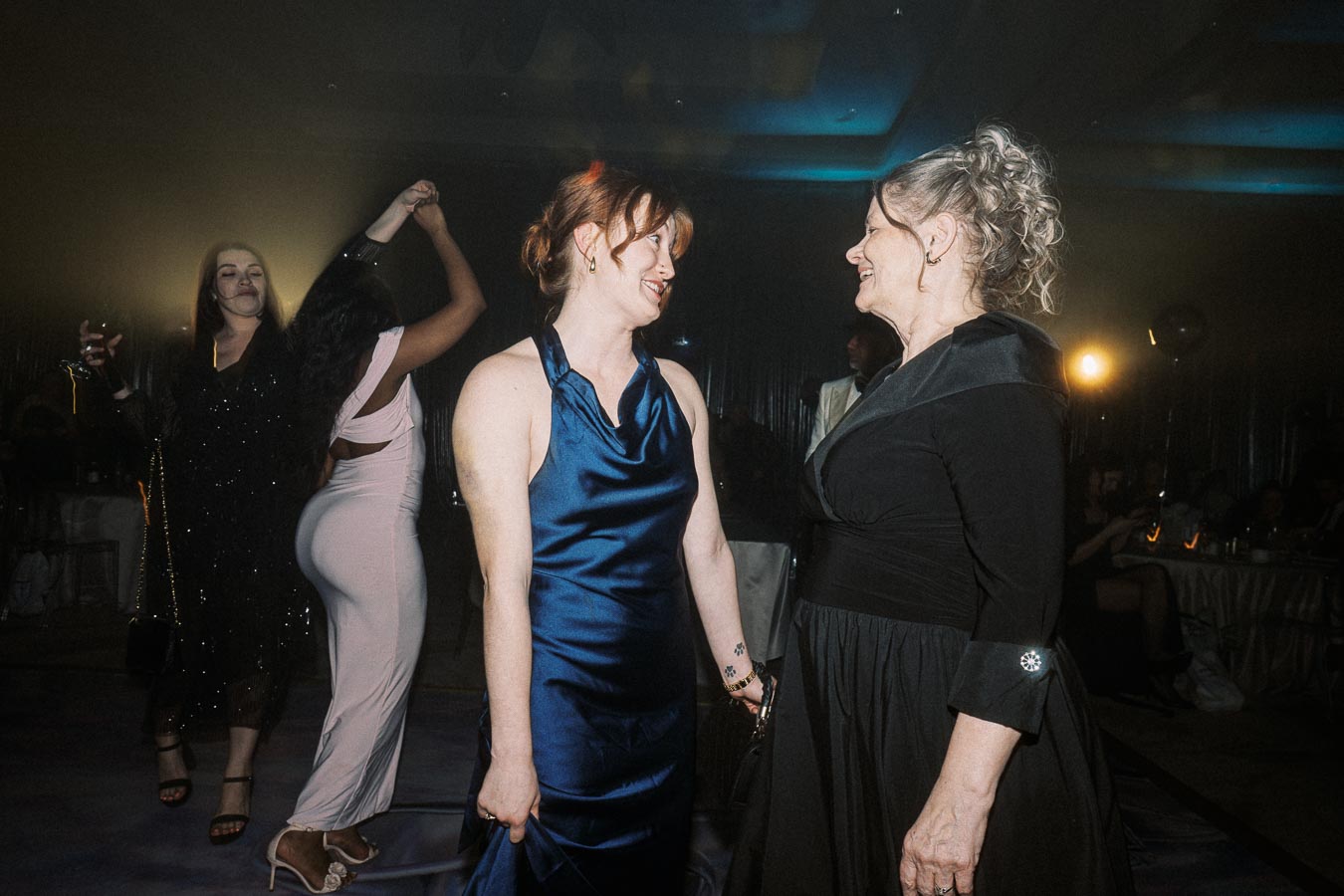 Women enjoying a night out at a party, dressed in elegant gowns, dancing and socializing in a dimly lit venue.