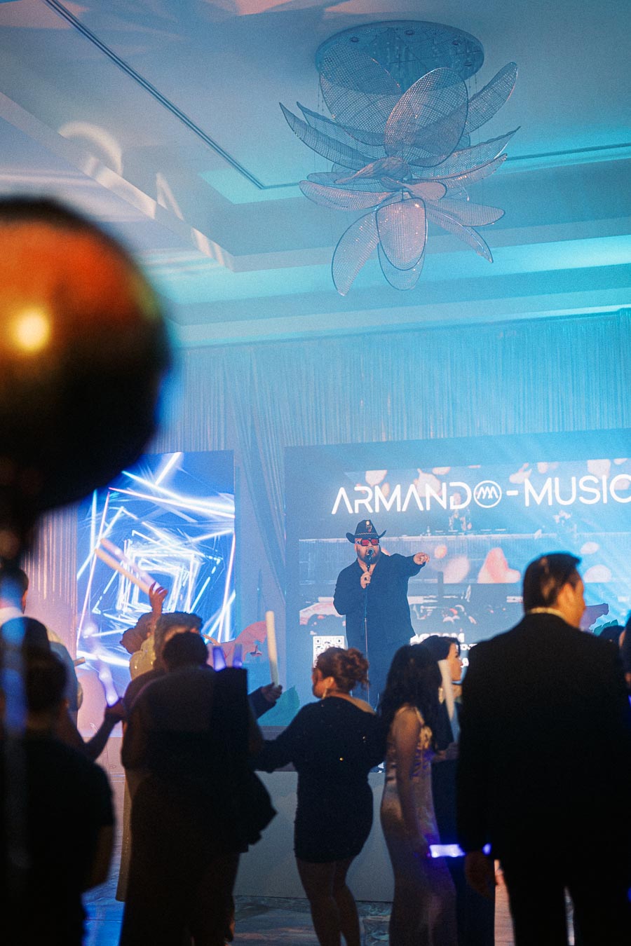 A lively indoor music event featuring a performer in a hat and sunglasses on stage with the words Armand Music in the background, surrounded by an enthusiastic crowd dancing and enjoying the concert under colorful lights.