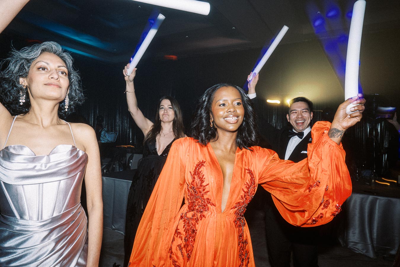 Elegant party scene with joyful guests dancing and holding glow sticks. A woman in a vibrant orange dress stands out, surrounded by others in formal attire, highlighting a festive and lively atmosphere.