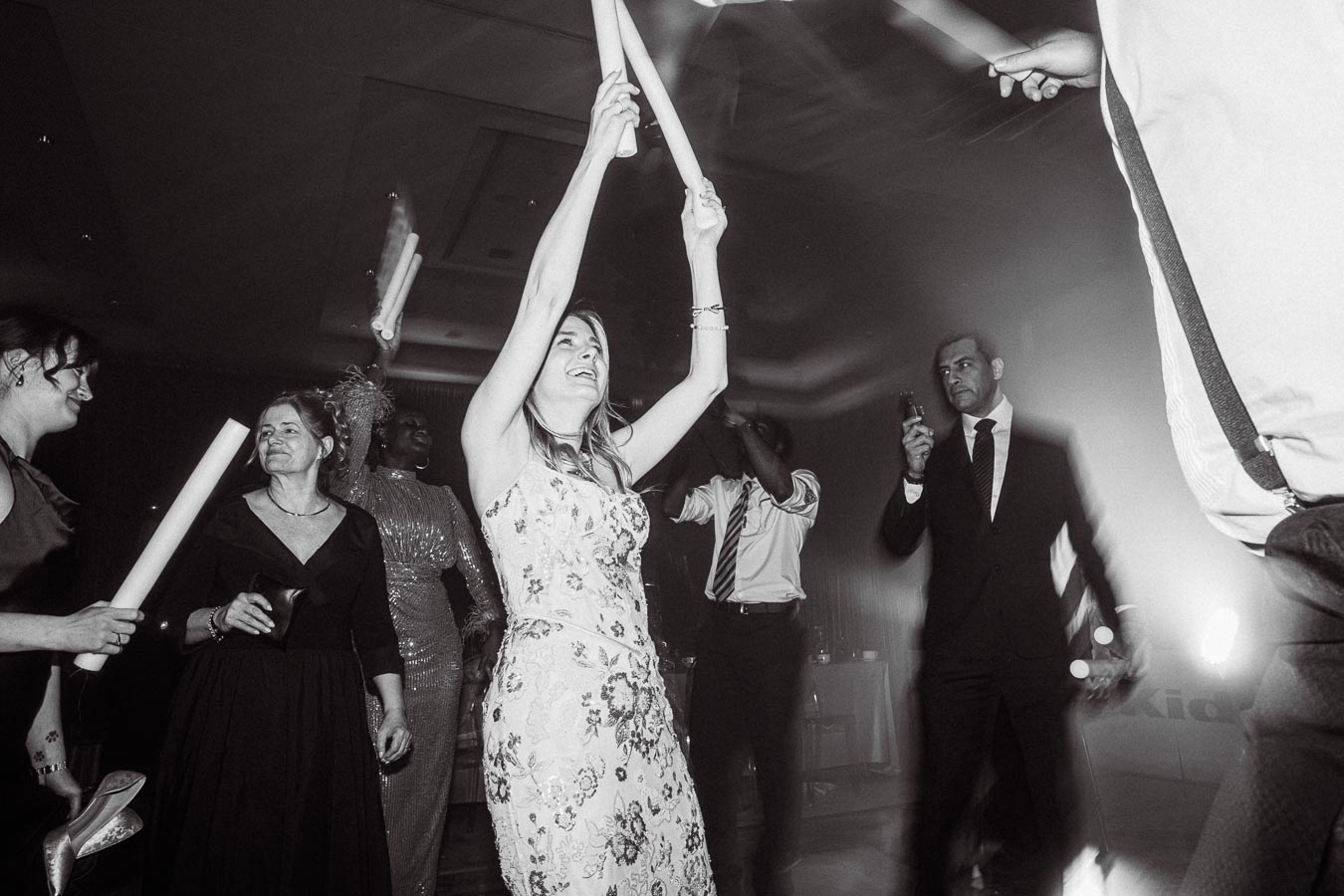 Black and white photo of a lively dance floor with elegantly dressed people, waving glow sticks and enjoying the celebration at a formal event or wedding reception.