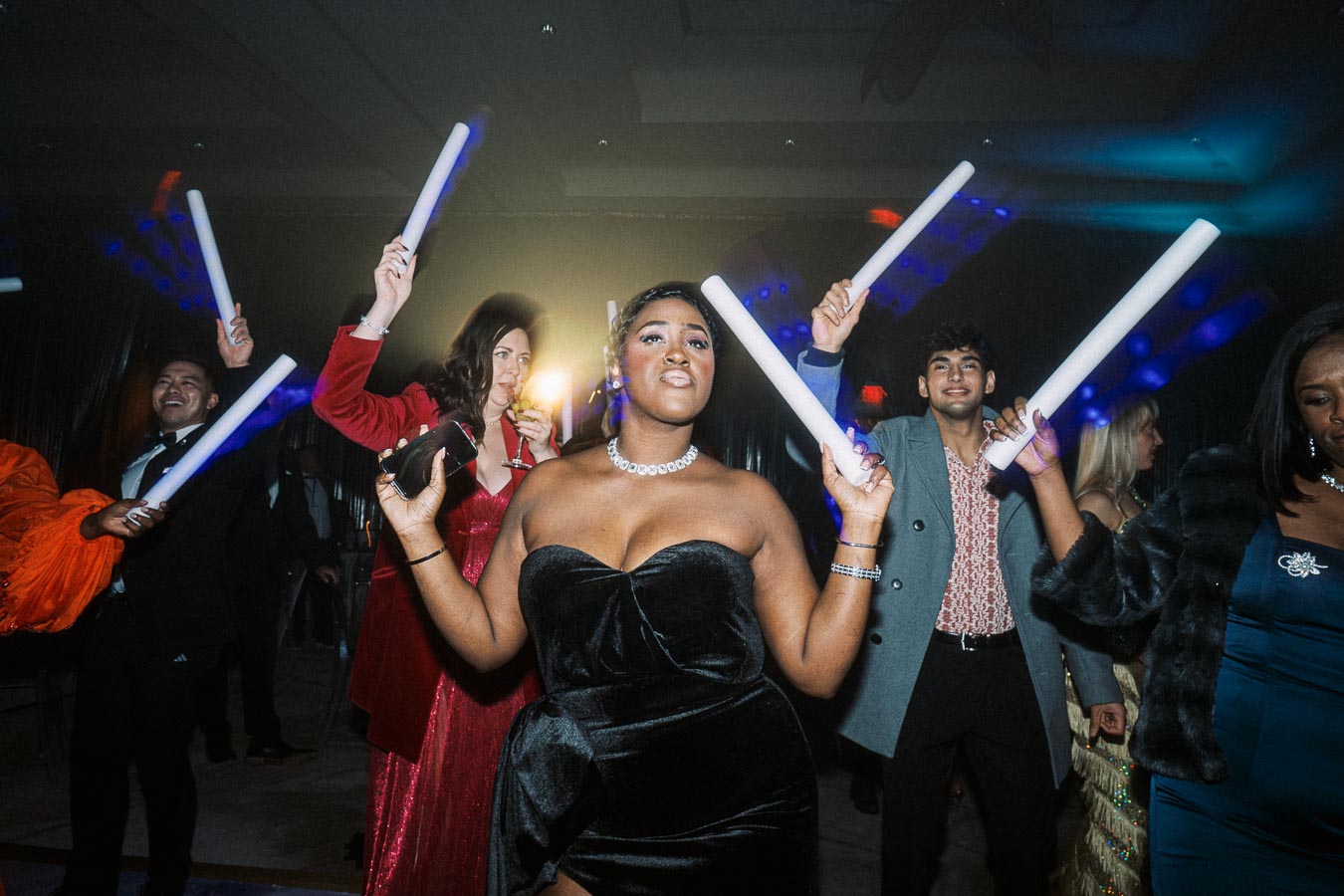 A group of people enjoying a lively party, dressed in formal evening attire while dancing and holding glowing light sticks, set against a dimly lit background with colorful lights.