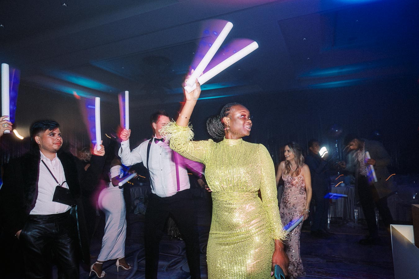 A lively party scene with elegantly dressed guests dancing and holding glowing sticks, featuring a woman in a sparkling green dress at the center. The ambient lighting creates a festive atmosphere, enhancing the celebration mood.