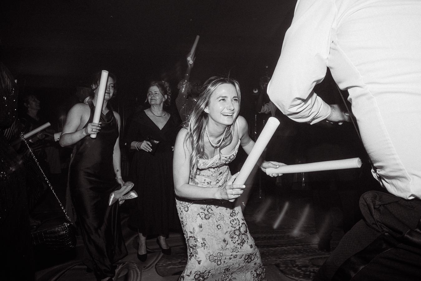 A lively black and white photo of a joyful party scene, featuring people dancing and smiling while holding inflatable sticks, with a woman in a floral dress in the foreground.