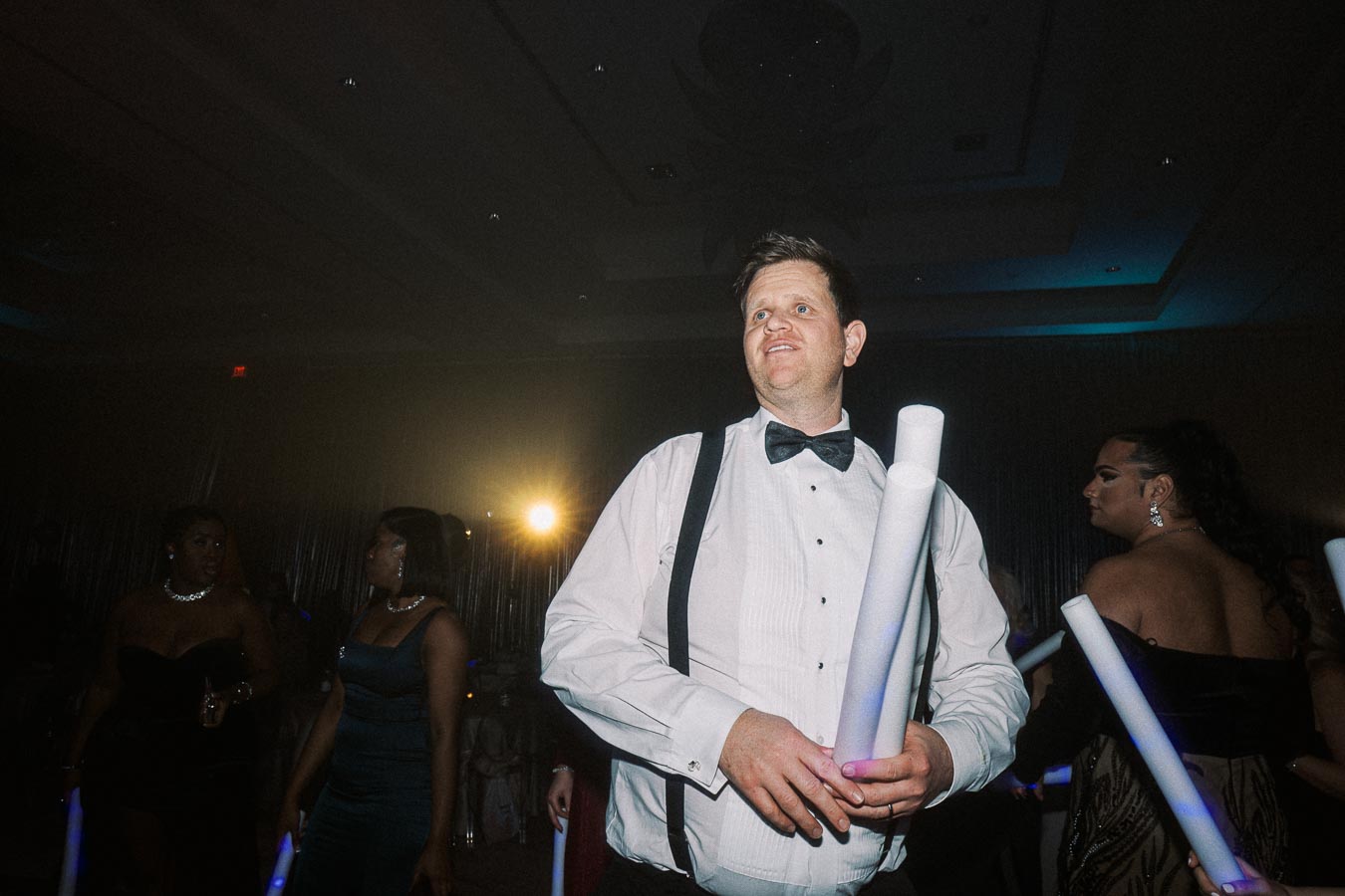 A man in a tuxedo with suspenders holding light sticks at a lively party, surrounded by elegantly dressed attendees, with a spotlight in the background creating a festive atmosphere.