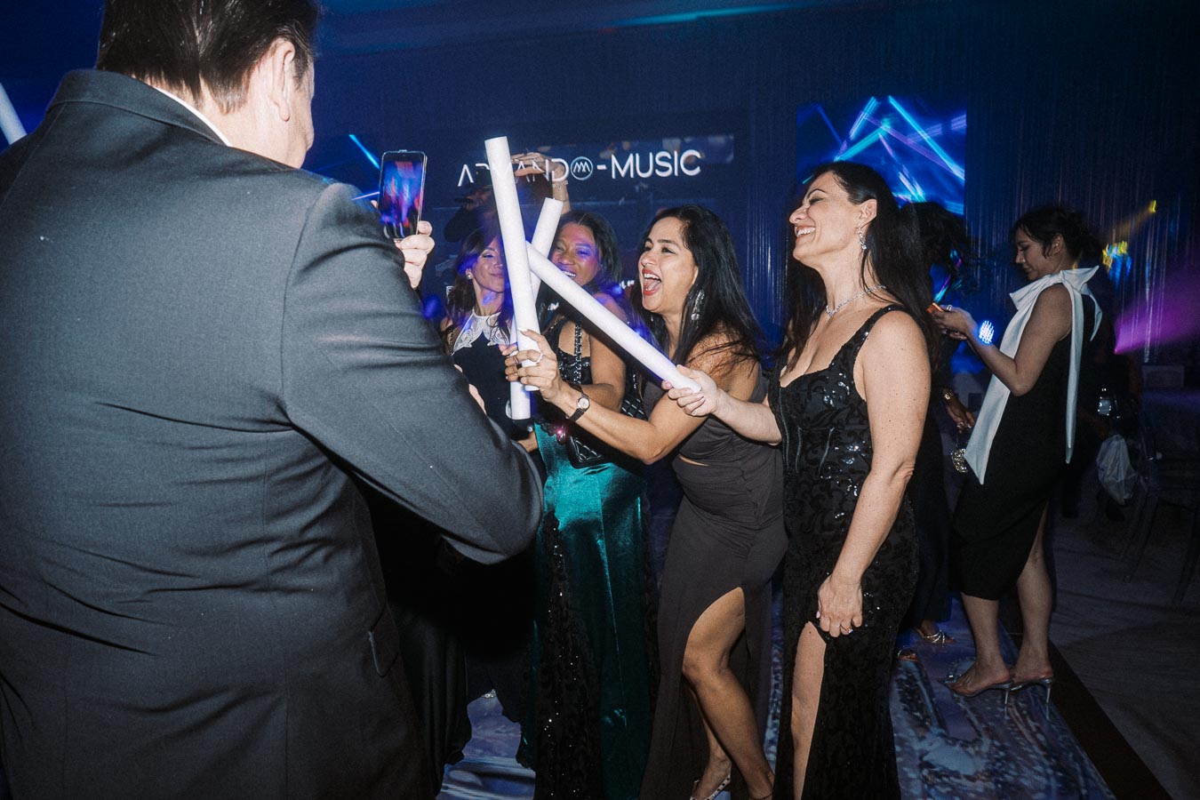 Group of people enjoying a lively party with music and dancing, holding light sticks and capturing moments on their smartphones.