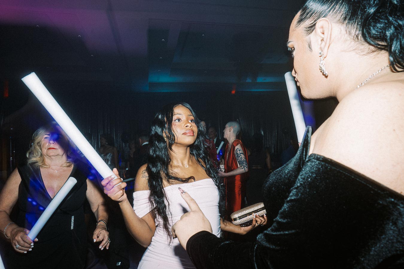 A lively party scene with elegantly dressed individuals holding glow sticks, surrounded by colorful lighting.