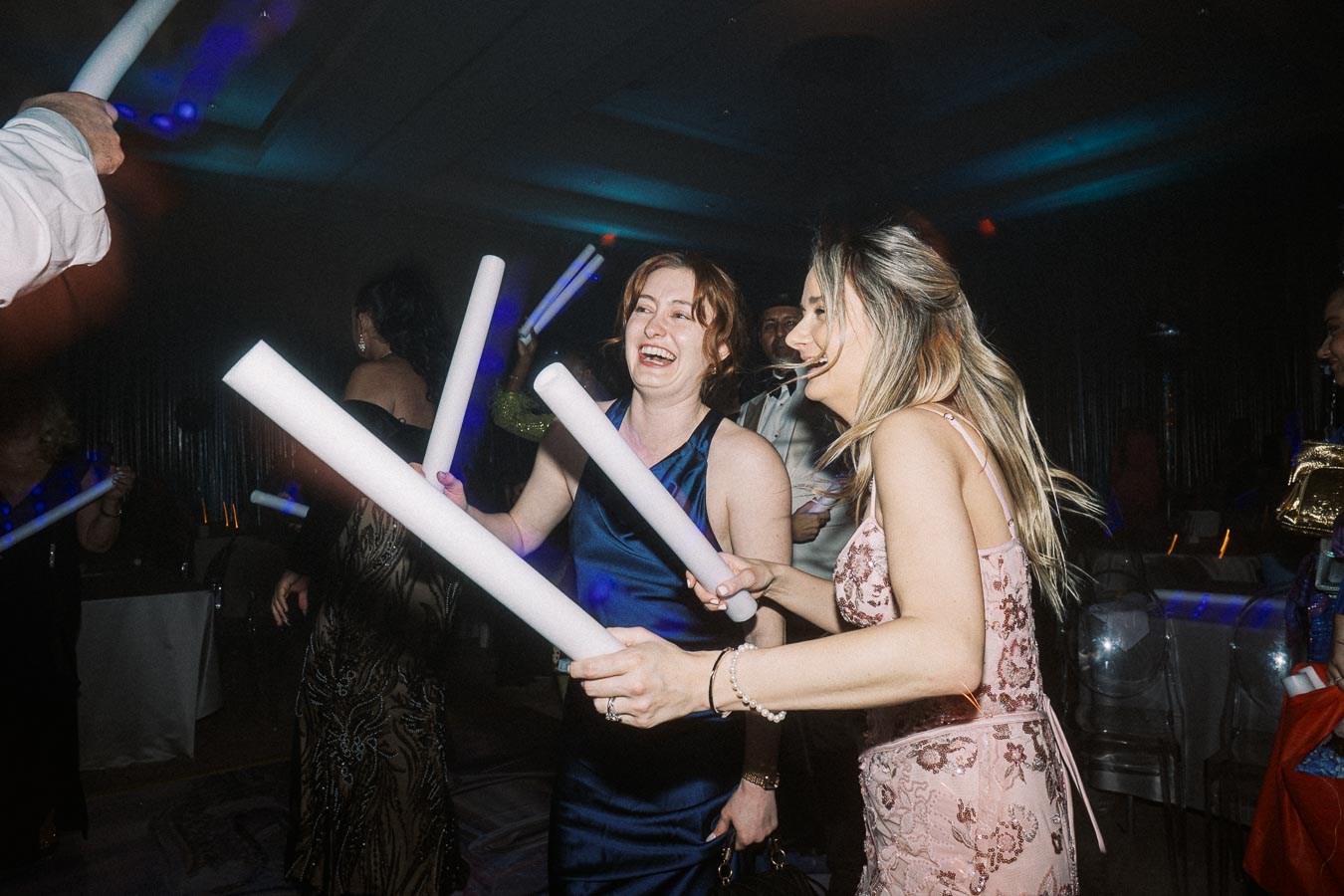 Women dancing at a lively party with glow sticks, dressed in elegant evening attire, enjoying the festive atmosphere under colorful lights.