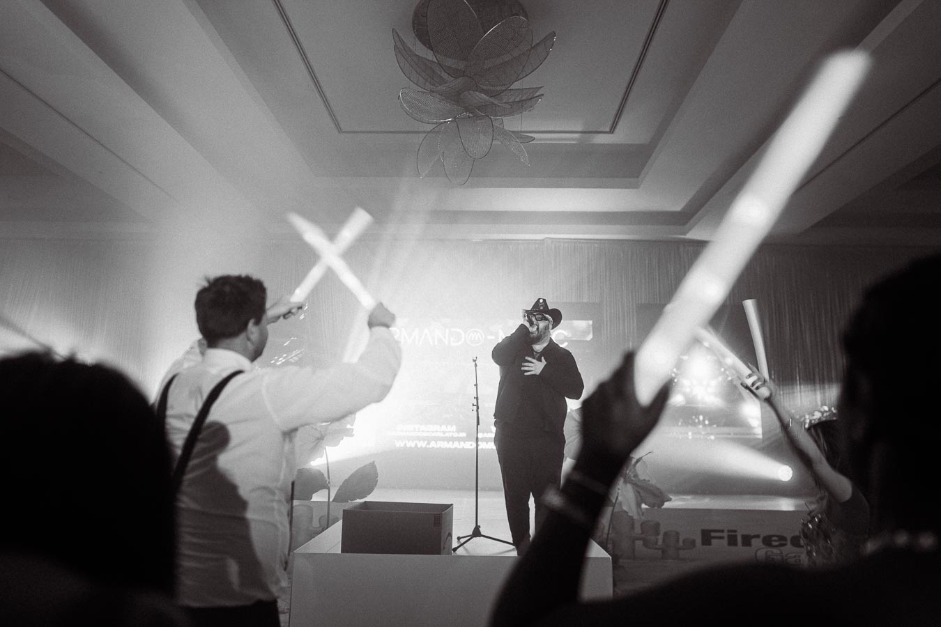 A performer in a cowboy hat sings passionately on stage at a lively concert, with fans energetically waving light sticks in the foreground.