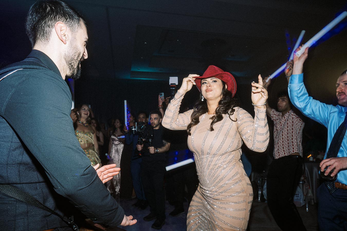 People dancing energetically at a party, with a woman in a red cowboy hat and sparkly dress leading the celebration, surrounded by guests holding glow sticks.