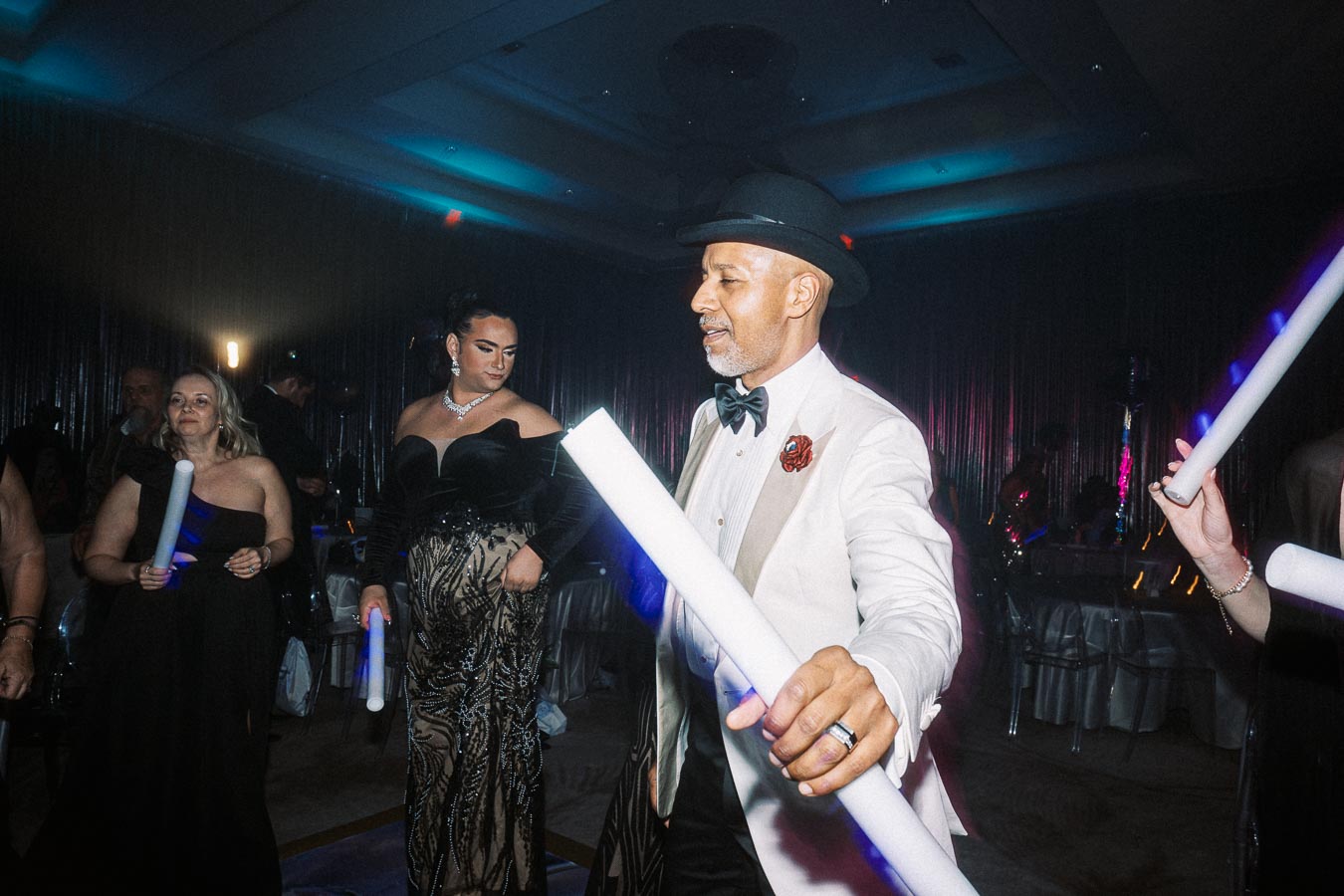 Elegant party scene with people dressed in formal attire holding light sticks, dancing in a dimly lit venue with ambient blue and purple lighting.