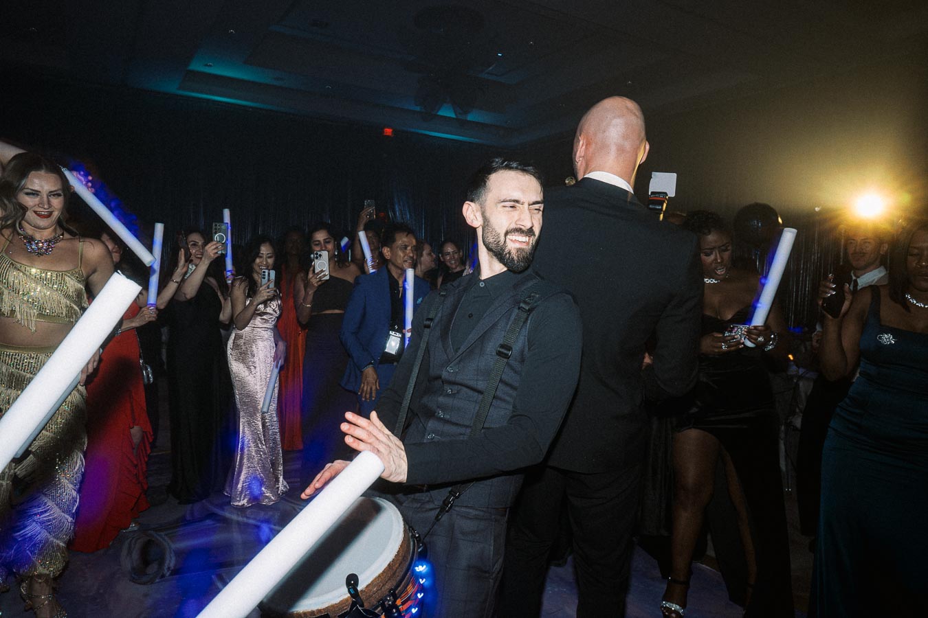 A lively party scene with a drummer energetically playing, surrounded by elegantly dressed guests holding glowing LED sticks and taking photos, creating a vibrant and festive atmosphere.