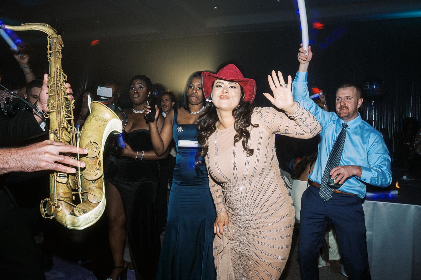 People dancing energetically at a lively party, with a saxophone player in the foreground. The scene includes a woman in a red hat and various others enjoying the vibrant atmosphere under party lights.