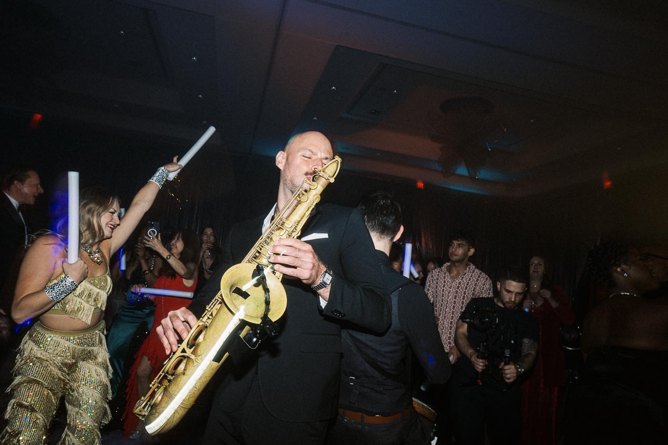 Man playing saxophone on a lively dance floor at a vibrant party, surrounded by elegantly dressed guests, dancing and celebrating under colorful lights.