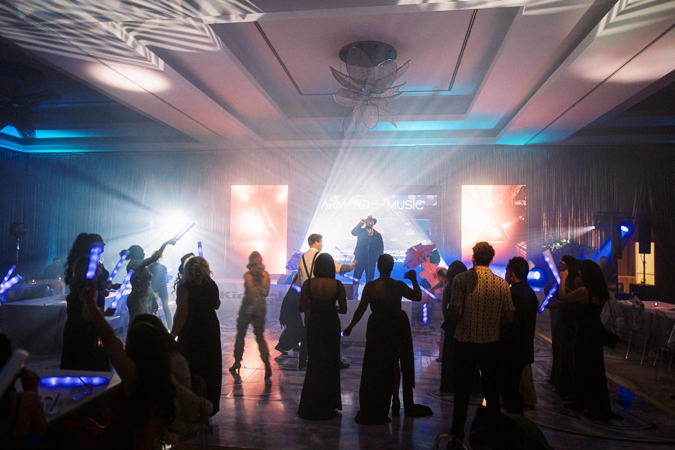 A lively dance floor scene at an indoor event with people celebrating under vibrant colored lights, while a DJ performs in front of large display screens.