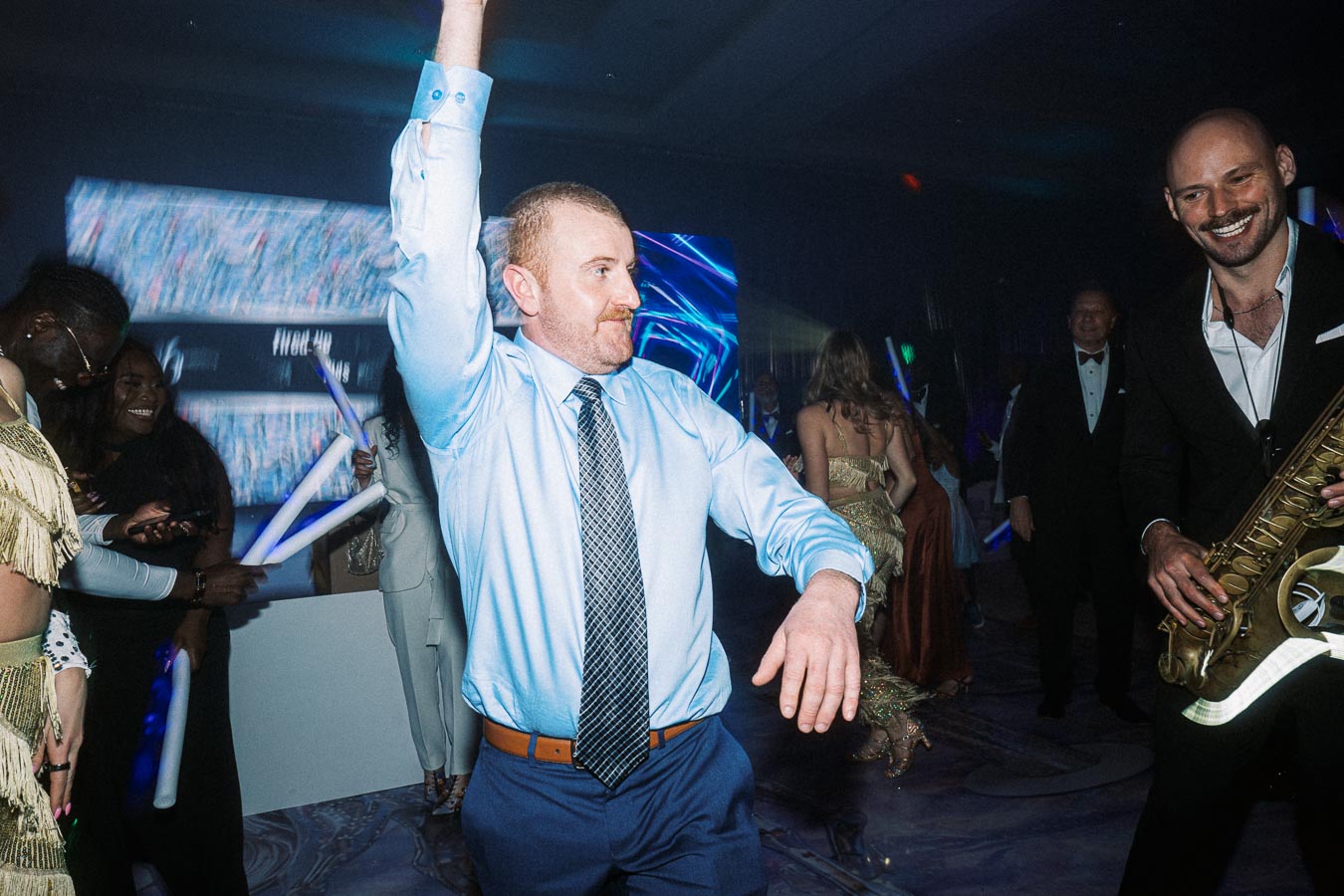 Man in a blue shirt enthusiastically dancing at a party with lively crowd and saxophonist in the background.