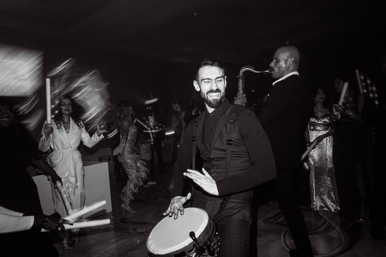 Musician playing a drum passionately at a lively party with guests dancing, some holding neon light sticks, and a saxophonist adding vibrant tunes in a dimly lit atmosphere.