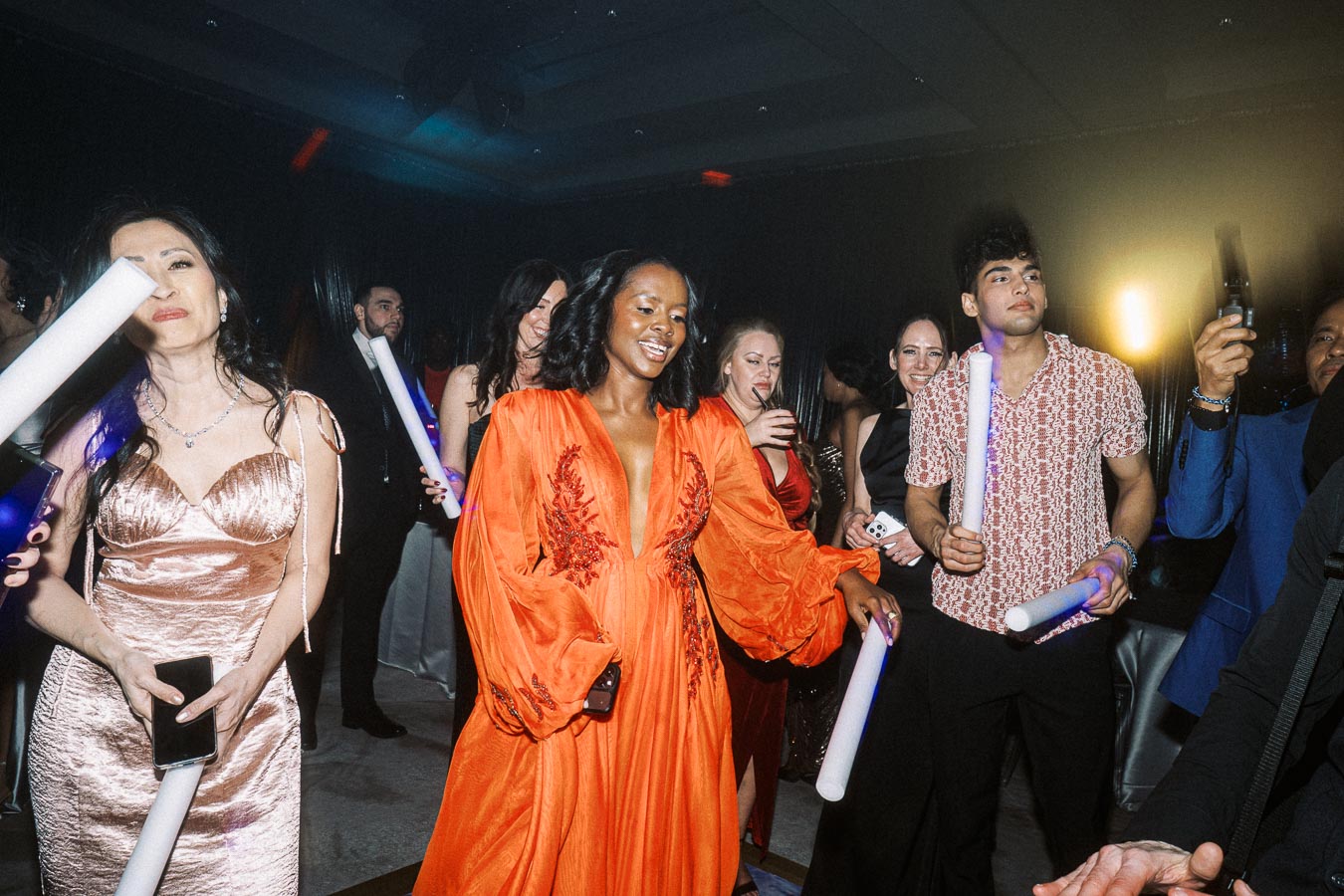 A lively party scene with a diverse group of people dancing and enjoying themselves, holding light sticks. A woman in an orange dress is at the center, surrounded by others in elegant attire, creating a vibrant and festive atmosphere.