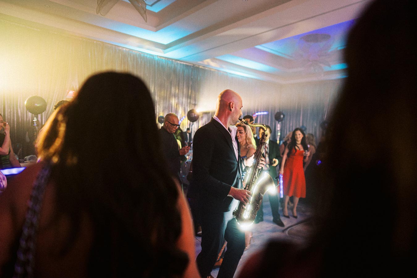 A lively indoor party scene with a saxophone player in a suit performing under colorful lighting, surrounded by elegantly dressed guests enjoying the music.