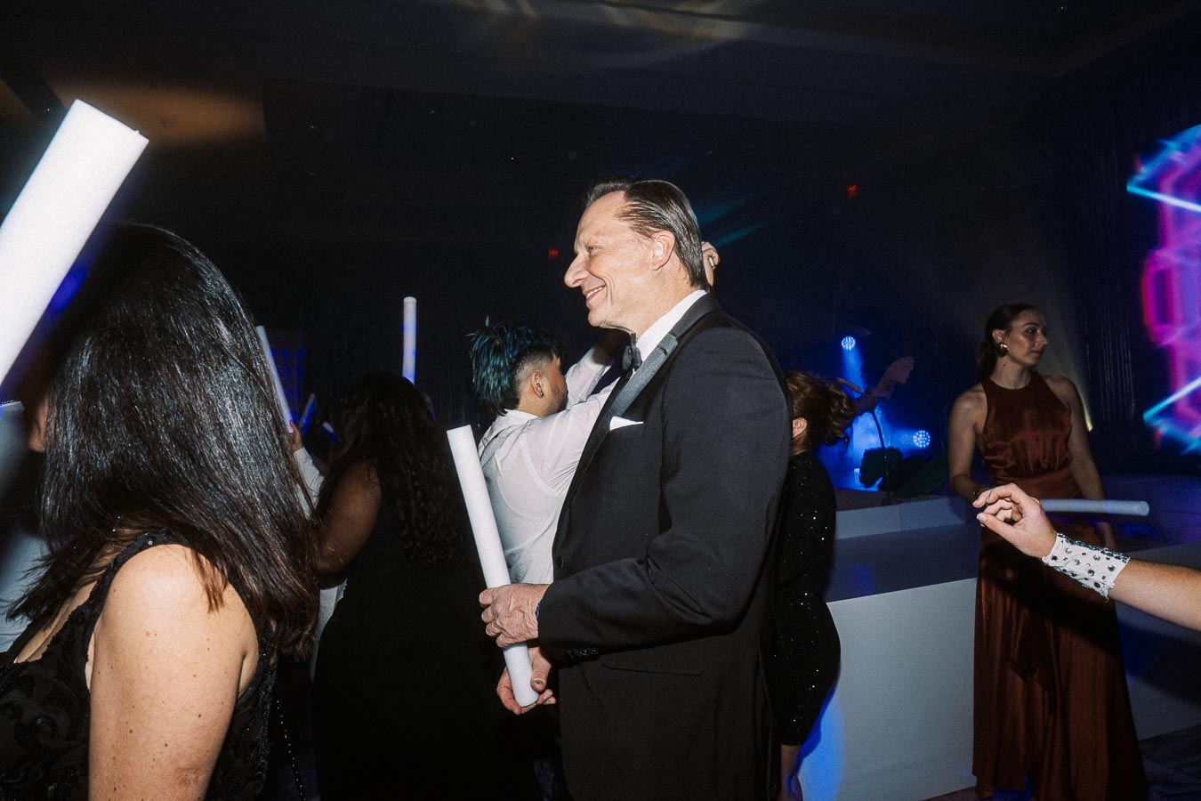 A lively party scene with elegantly dressed guests dancing under colorful lighting. The focus is on a smiling man in a tuxedo holding a light stick, surrounded by other attendees in formal attire enjoying the vibrant atmosphere.