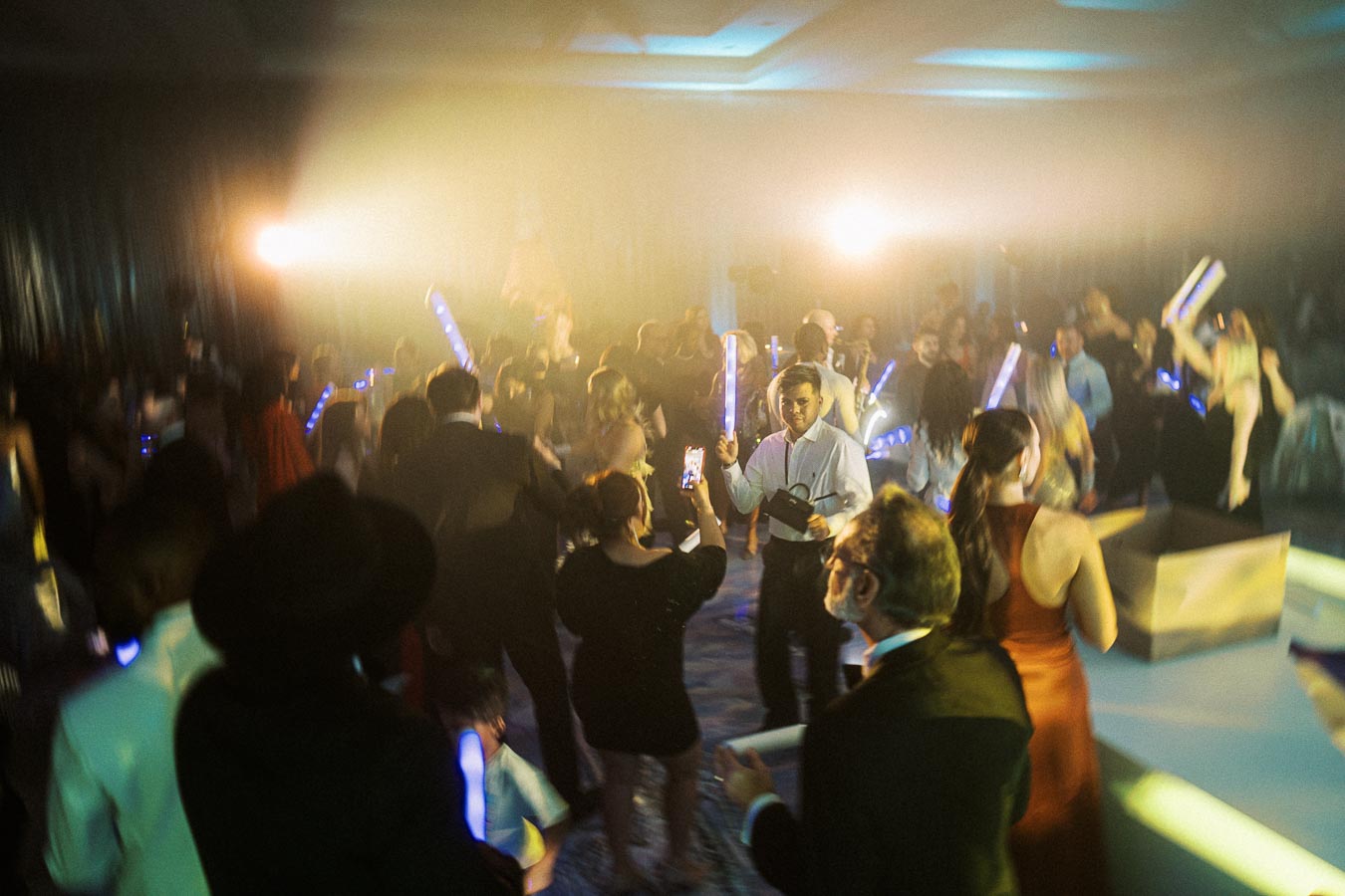A vibrant party scene with a crowd of people dancing under colorful lights. The atmosphere is lively, filled with guests dressed in formal attire and waving illuminated sticks. A sense of celebration is enhanced by multicolored lighting and hazy effects, creating an exciting and festive mood.