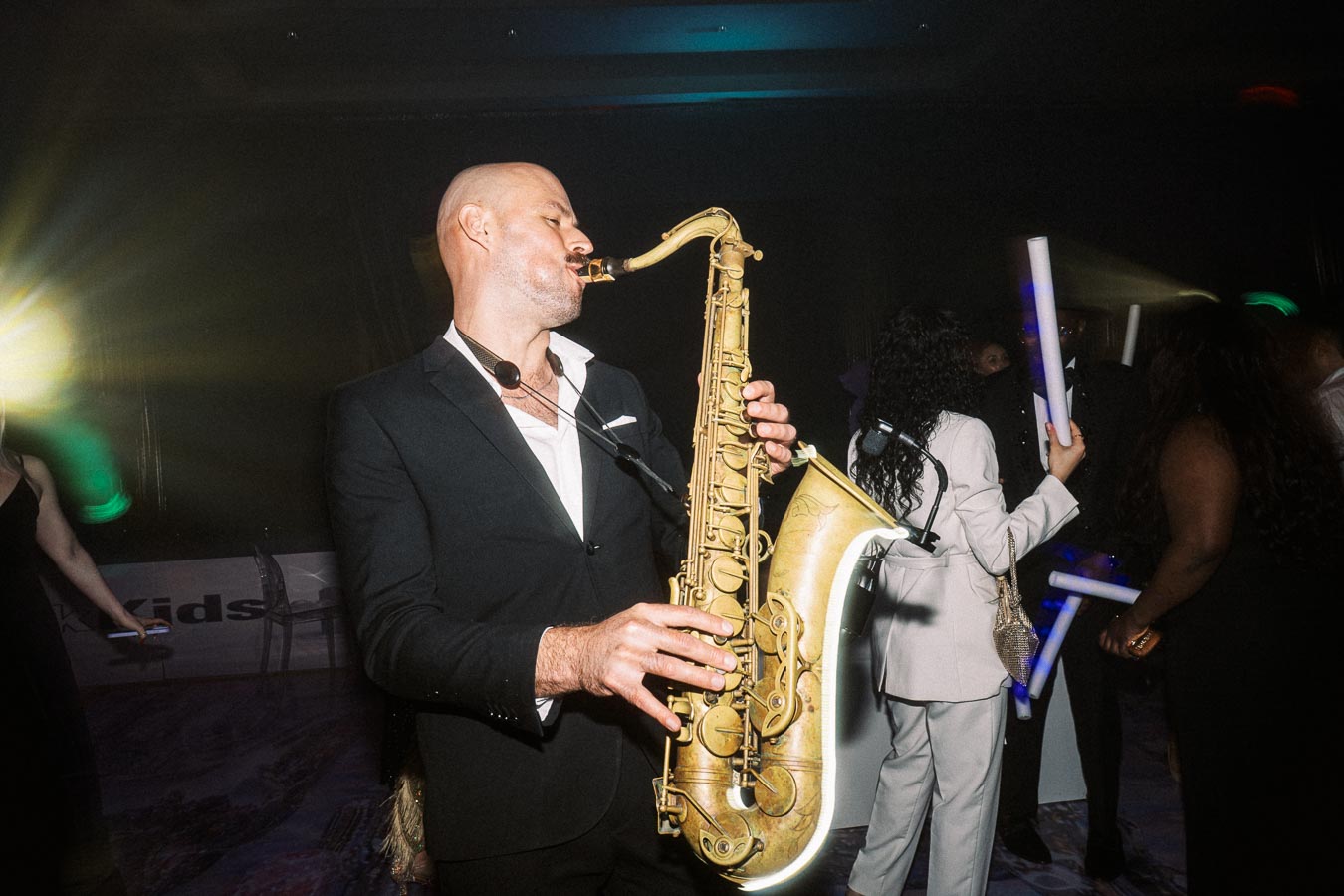 Saxophonist in a black suit passionately playing at a lively party event with attendees dancing and vibrant lighting in the background.