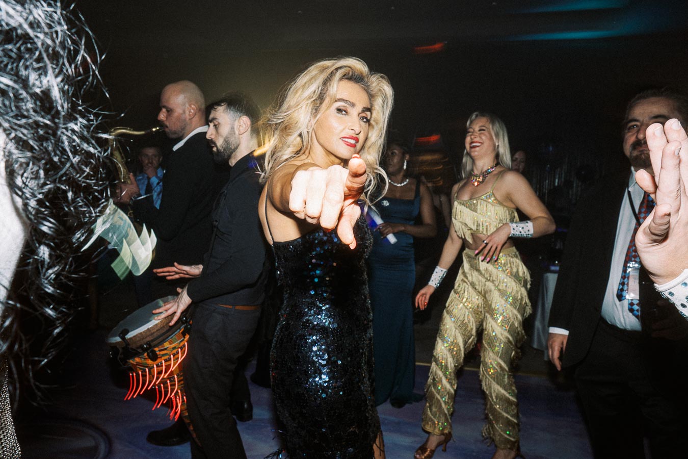 A lively party scene with a woman in a sequined dress pointing towards the camera, surrounded by people dancing and musicians playing instruments, creating a vibrant and energetic atmosphere.