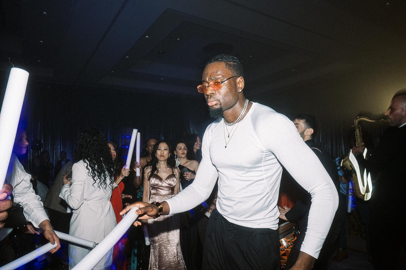 A lively party scene with a man in a white shirt holding a light stick, surrounded by other partygoers in festive attire, enjoying the atmosphere with live music in the background.