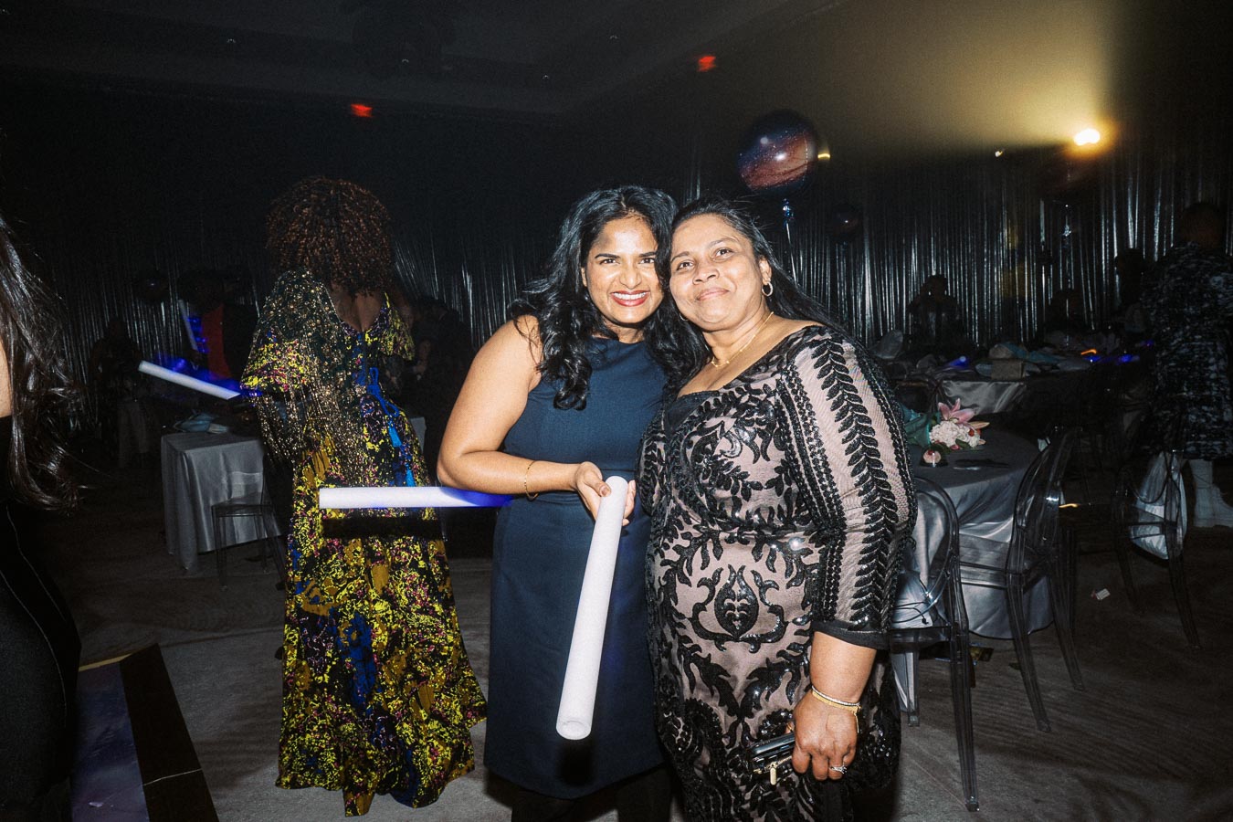 Two women smiling and posing with glow sticks at a lively party, dressed in elegant evening gowns, with a dimly lit background showcasing a decorated event space.