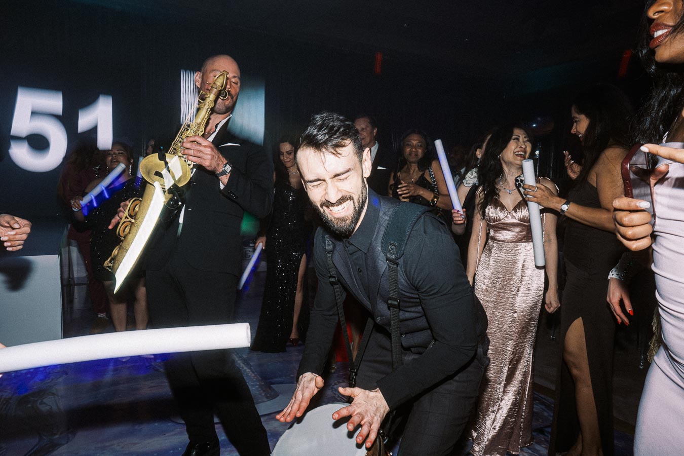 A lively party scene with a man passionately playing the saxophone while guests in elegant attire dance and enjoy the music, holding glowing sticks.